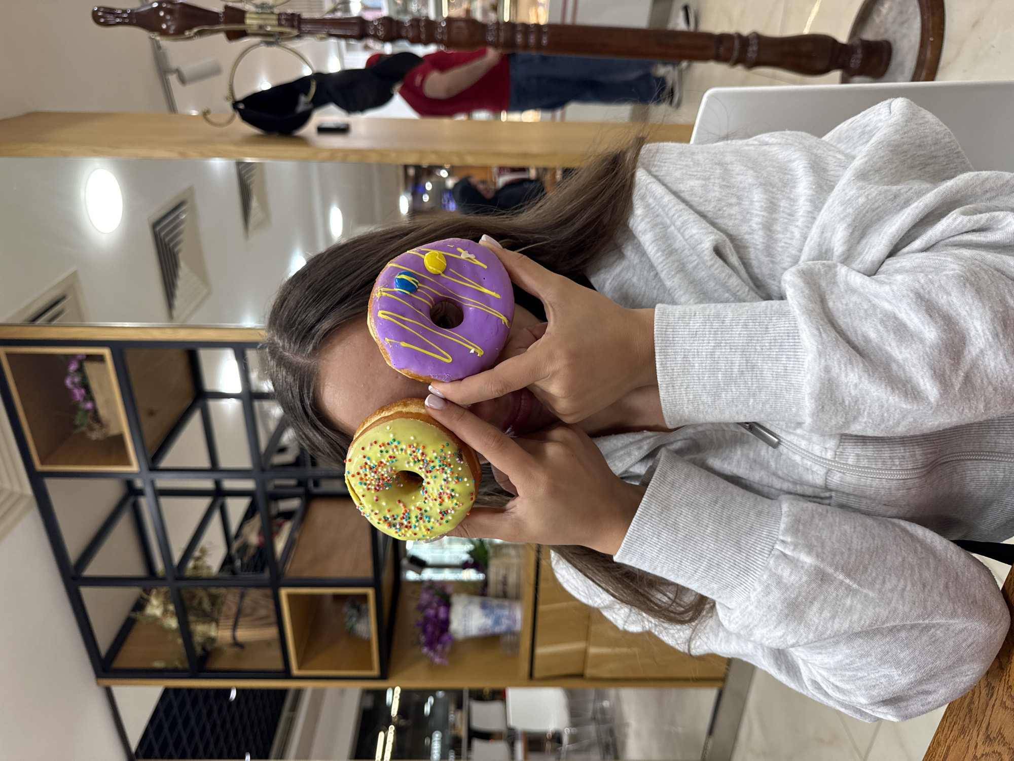 A young woman is holding two donuts in front of her face. The top donut is purple with yellow icing and yellow and blue sprinkles. The bottom donut is green with colorful sprinkles. The woman is wearing a grey hooded sweatshirt. A mirror behind her reflects a person in a red shirt. A wooden display shelf is visible on the left, and a decorative wooden pole is visible at the top of the image. The location is inside a donut shop, but the city of Aparan, Armenia cannot be determined from the image.