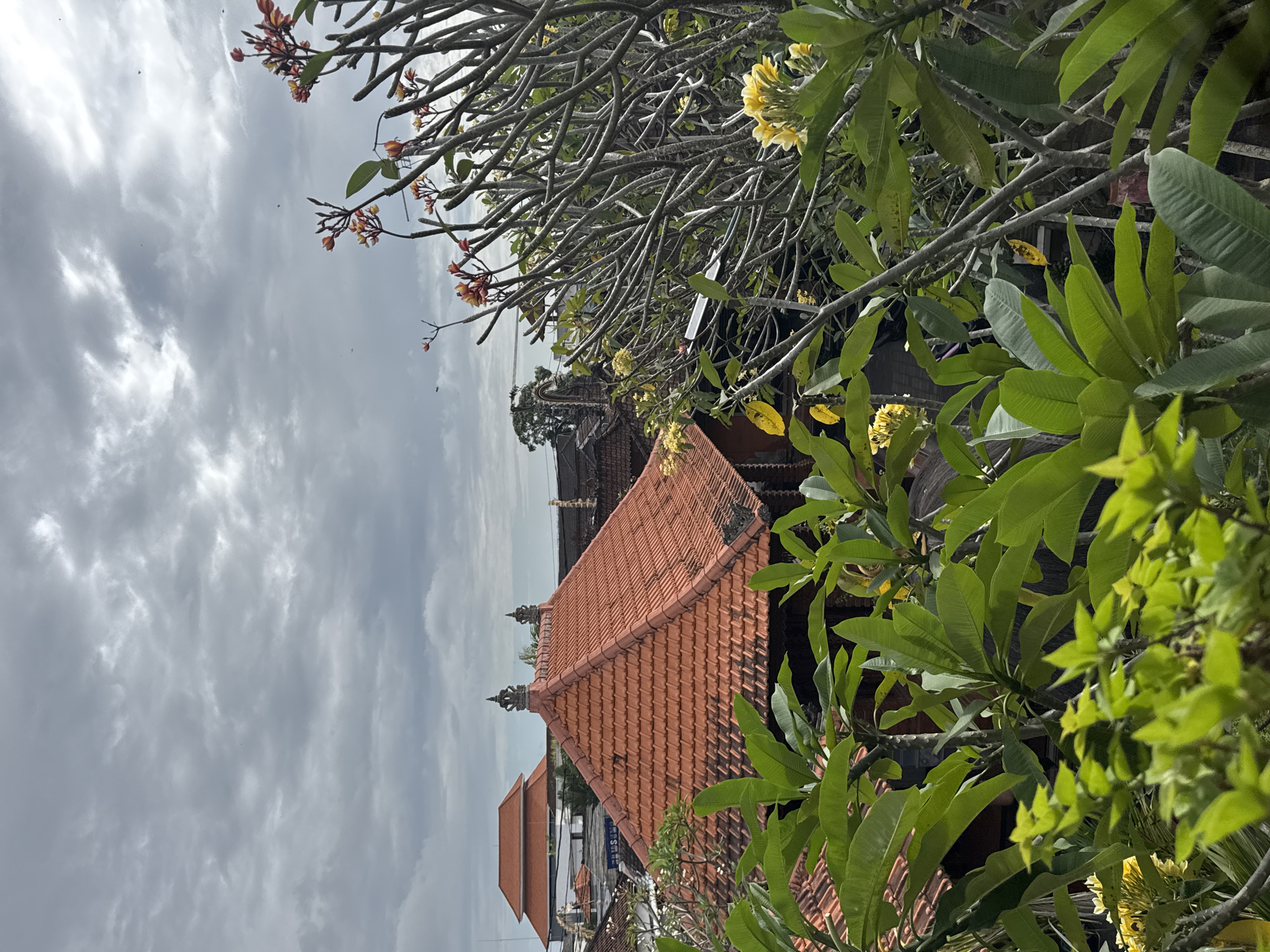 An overcast sky looms above a cluster of buildings in North Kuta, Indonesia. In the foreground, lush green foliage, likely from a plumeria tree, partially obscures a traditional red-tiled roof, a common architectural feature in the region. The roof is peaked and adorned with decorative finials. Beyond this, other buildings with similar red roofs are visible, suggesting a dense urban or semi-urban environment. The scene is quiet, with no people or discernible activity. The overall impression is one of tropical architecture under a moody sky.