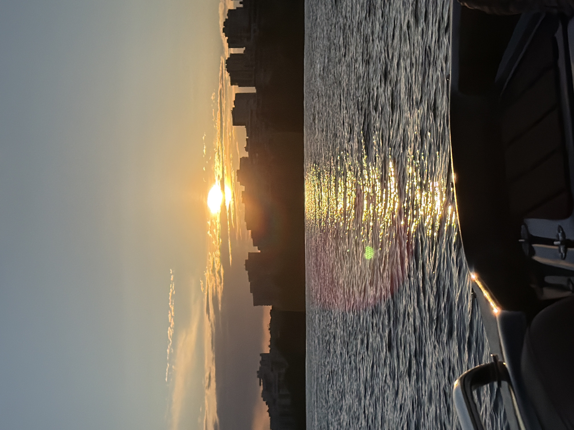 This is a low-angle, medium shot of a boat deck at sunset, with the Kazan skyline visible in the background. The sun is setting behind a line of silhouetted buildings on the horizon, casting a golden glow on the water. The sky is a gradient of blue and orange, with some scattered clouds. The water is choppy, with reflections of the sun rippling across its surface. The boat deck in the foreground is dark and appears to be made of leather or vinyl, with a chrome handle visible on the left side. The scene is tranquil and picturesque, evoking a sense of calm and relaxation.