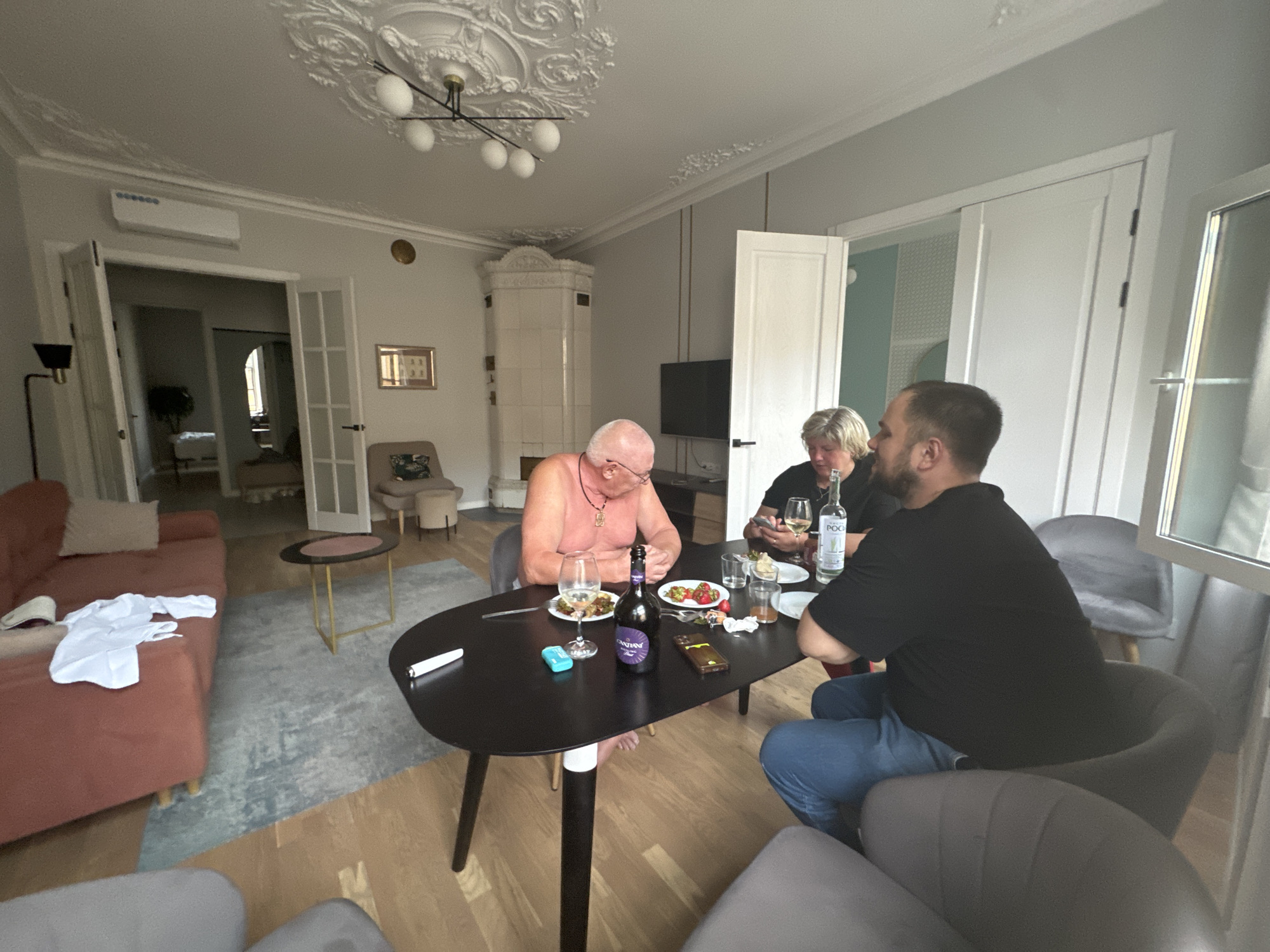 The image shows an interior scene in what appears to be a modern apartment in Saint Petersburg, Russia, likely a living or dining area. Three people are seated around a black, oval dining table. A man with grey hair and glasses is wearing a pink shirt, and he is holding a glass. To his right is a woman with blonde hair wearing a black top, and she is looking at her phone. The third person, a man with dark hair, is wearing a black t-shirt and blue jeans, and he is also seated at the table, looking down. On the table are plates of food, glasses, a bottle of wine, and a bottle of water with the label "POCHI."
The room features a light-colored rug on a wooden floor, a coral-colored sofa with a white garment on it, and two modern armchairs in grey. In the background, there are doorways leading to other rooms, a white tiled oven or fireplace, and a television mounted on the wall. The ceiling has ornate plaster molding and a modern chandelier. The overall impression is of a casual gathering or meal in a well-decorated living space.