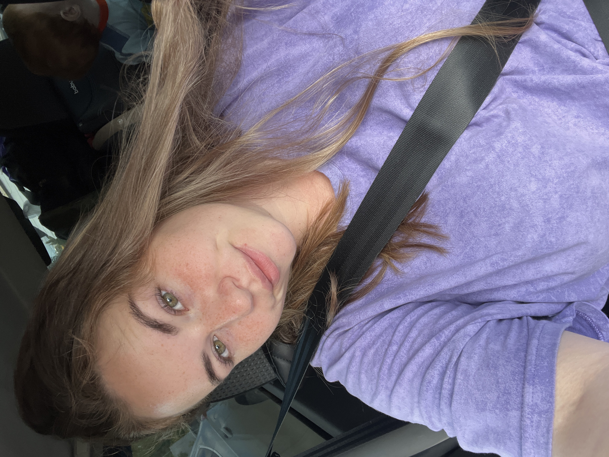 A young woman with light brown hair and green eyes is taking a selfie in a car. She is wearing a purple t-shirt and a black seatbelt. Her hair is long and wavy, and she has freckles on her face. In the background, a child can be seen in a car seat. The image appears to be taken in daylight. There is no indication of the location being in Chaikovsky, Russia.