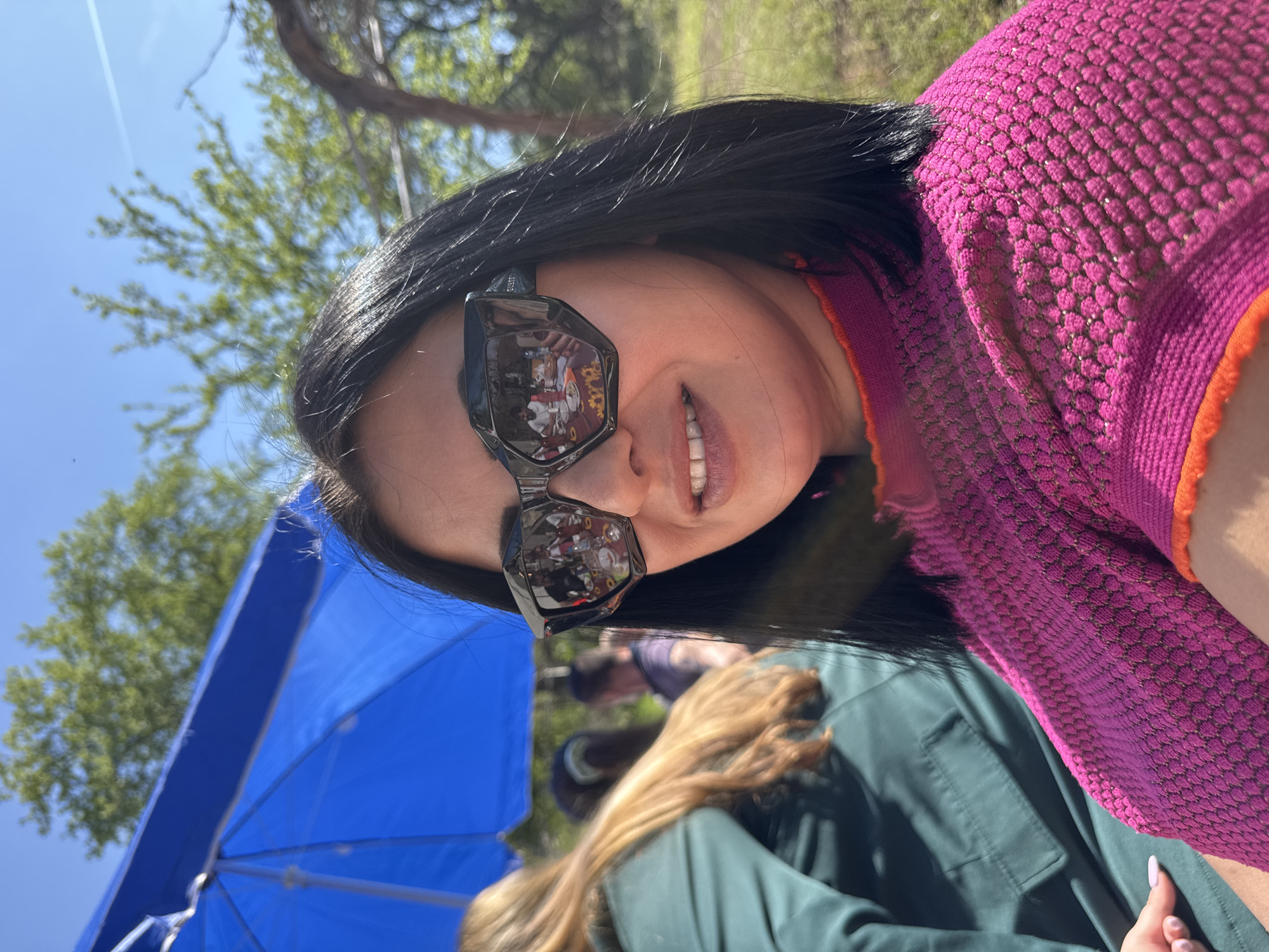 A woman with dark hair wearing dark sunglasses and a pink sweater is photographed outdoors. The location appears to be a social gathering, as people are visible in the background and reflected in her sunglasses. A large blue umbrella is also visible. While the precise location in Lom, Bulgaria, is not directly identifiable from the image, the scene suggests an open-air public space, perhaps a park or event venue.