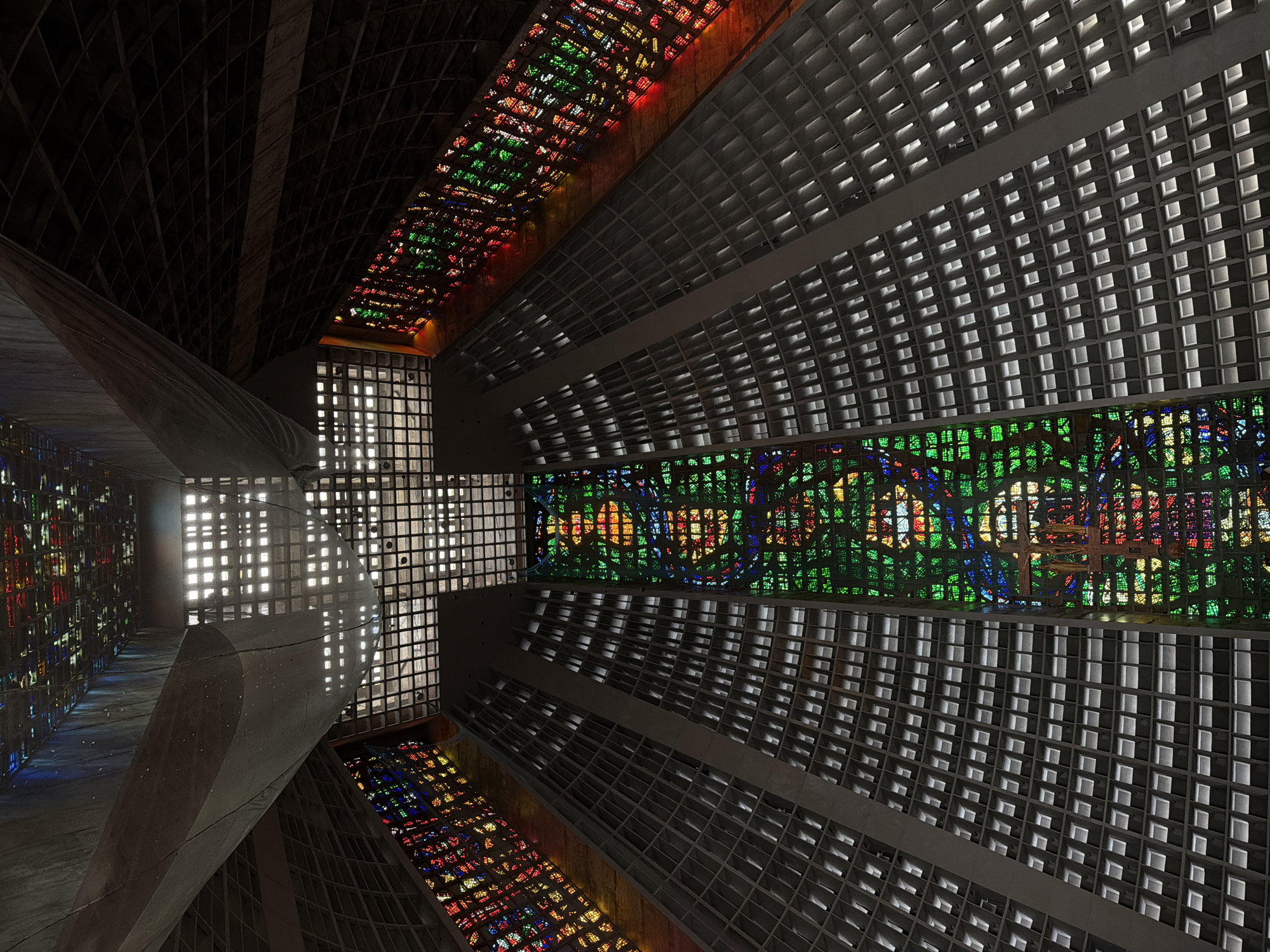 The image shows the interior of the Metropolitan Cathedral of Saint Sebastian of Rio de Janeiro, Brazil. The camera is looking upwards from the floor of the nave towards the ceiling. The architecture is characterized by a conical shape with four large parabolic arches that meet at the apex, where a massive stained-glass cross is situated.
The walls of the cathedral are lined with a grid of rectangular windows, many of which are illuminated from within, creating a striking pattern of light and shadow. On either side of the nave, extensive stained-glass panels run vertically, flooding the space with vibrant colors. The central stained-glass panel, directly above the altar area, is predominantly green and features a depiction of a crucifix with winged figures.
The ceiling features a complex grid structure, with some sections allowing light to filter through, creating a diffused glow. A large, fabric-like material hangs from the ceiling in the upper left portion of the image, casting shadows. The overall impression is one of grandeur, modernity, and the dramatic interplay of light and color through the architectural design and stained glass. There are no people visible in the image.