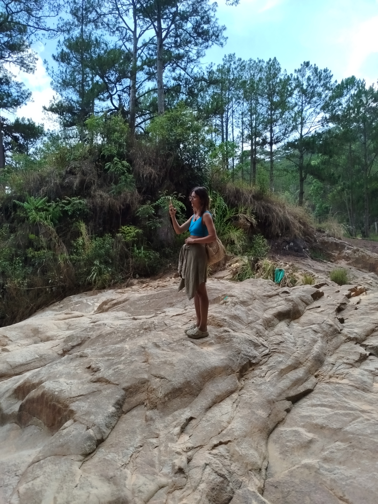 A woman stands on a large, smooth rock formation in a forested area, possibly a riverbed, in Đa Thọ, Vietnam. She is fair-skinned, wearing a blue top, a tan skirt, and shoes. She has sunglasses on her head and is holding a phone up as if taking a photo. Behind her is a lush, green hillside with trees. The sky is overcast.