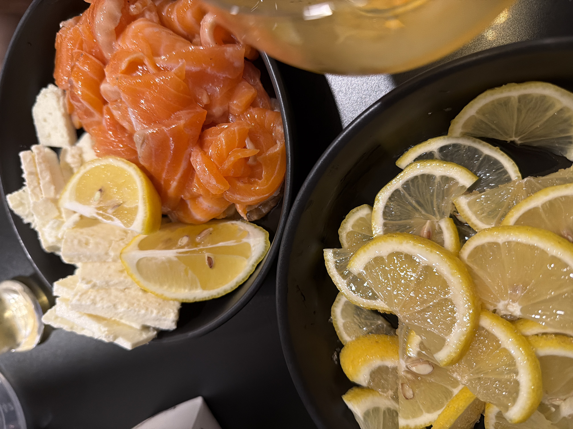 Two black bowls are positioned on a dark surface. The bowl on the left contains sliced salmon, rectangular pieces of white cheese, and lemon wedges. The bowl on the right is filled with numerous lemon slices. A clear liquid is being poured from a container onto the ingredients. The location is Kathu, Thailand.