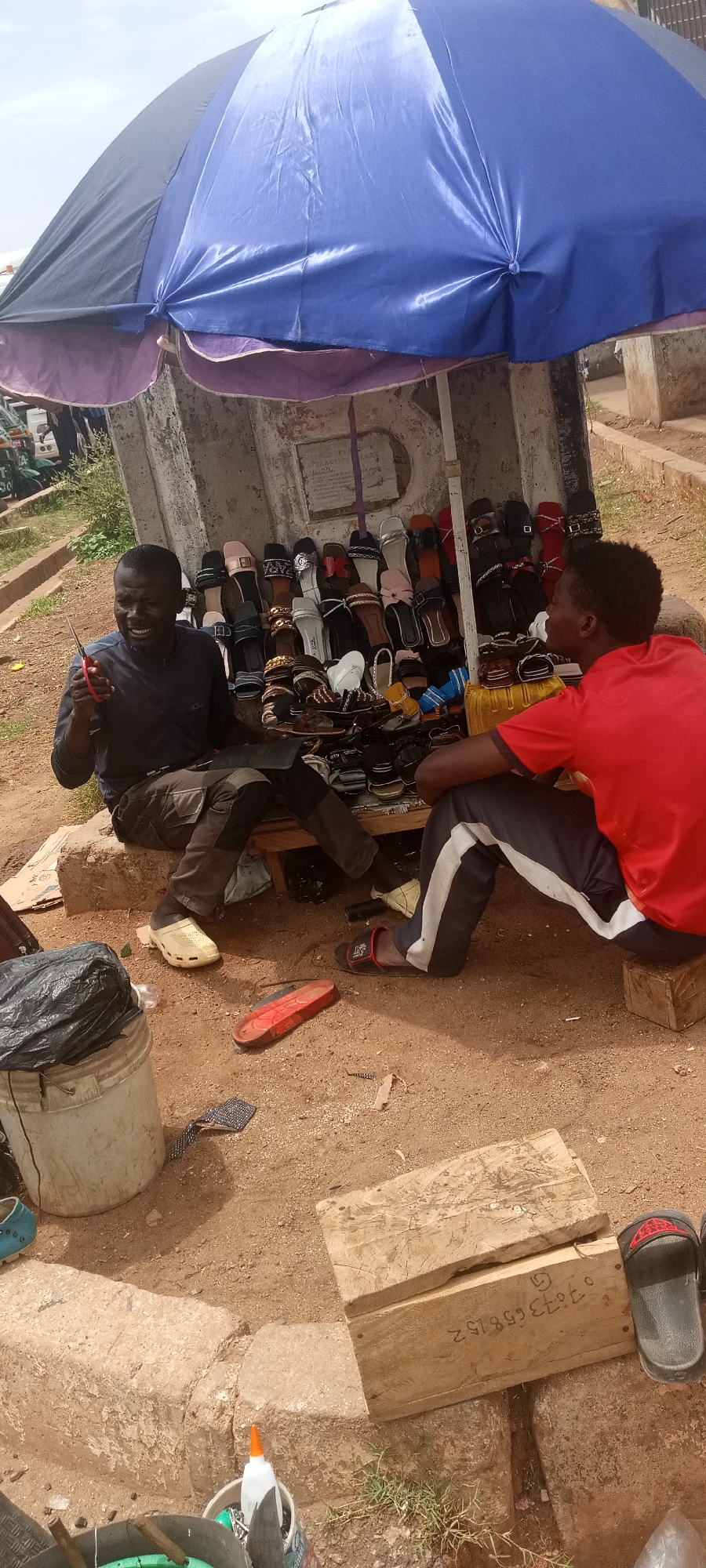 The image shows a street vendor selling sandals in Gura Top, Nigeria. Two men are present. One man sits behind a display of sandals under a blue umbrella. He holds scissors and appears to be interacting with a customer sitting to his right, who is wearing a red shirt. The ground is dirt, and there are various objects scattered around, including a bucket, a piece of red sandal, and a wooden box with writing on it. The background shows some vegetation and parked vehicles. The sandals are of various designs and colors.