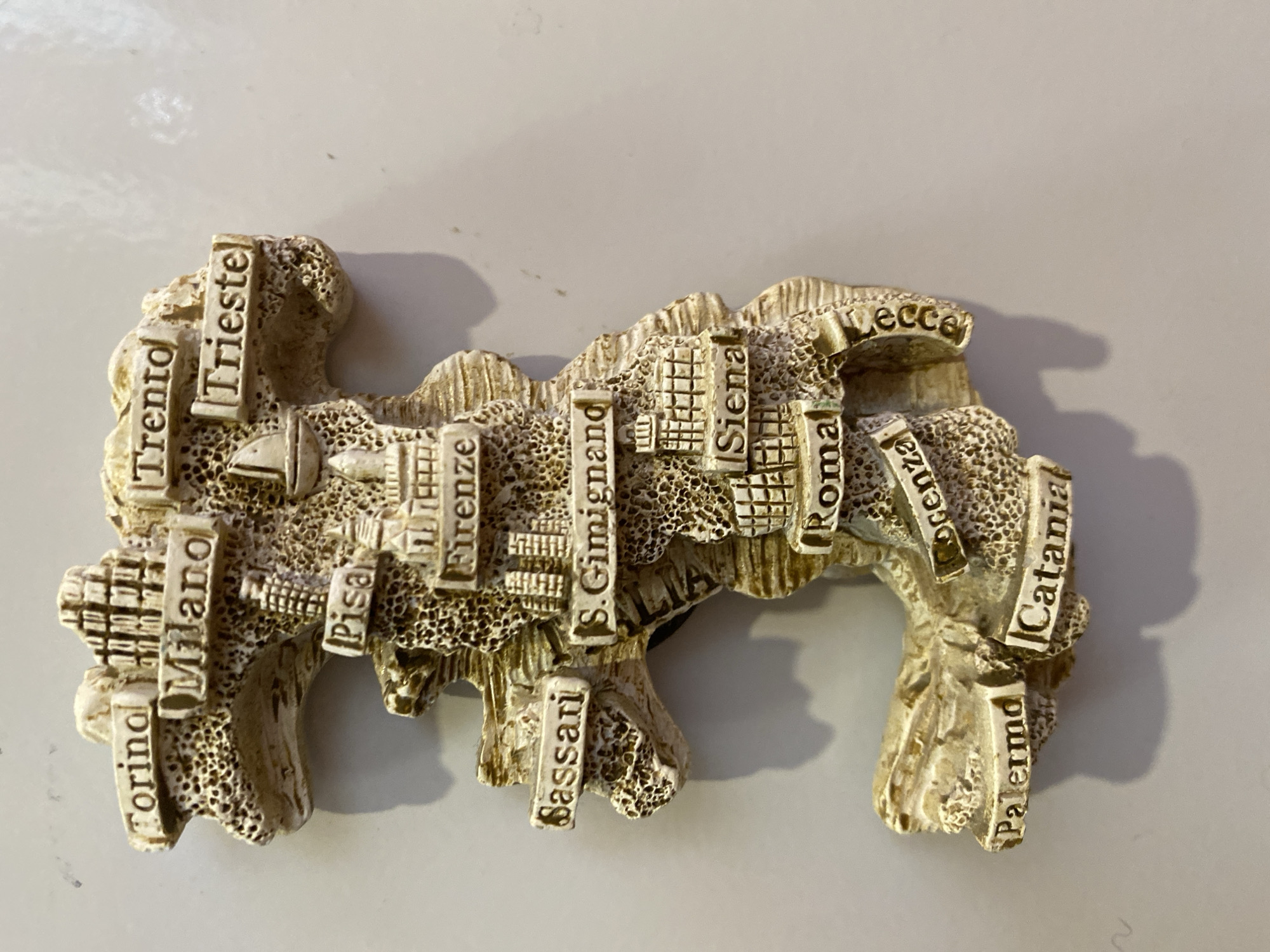This is a close-up, overhead shot of a decorative, three-dimensional map of Italy. The map is made of a light-colored, textured material, possibly resin or ceramic, and has a distressed, antique appearance. It is shaped like the boot of Italy, with various cities embossed on it. These cities are labeled with text in dark capital letters.
From left to right, the visible cities are: "Torino", "Milano", "Trento", "Trieste", "Pisa", "Firenze", "S. Gimignand" (likely San Gimignano), "Siena", "Roma", "Puglia" (partially obscured, appears as "ecce"), "Cosenza" (partially obscured, appears as "Ocenza"), "Palermo", and "Catania". The surface of the map is uneven and pitted, simulating a rocky or ancient texture.
The object is placed on a plain, light-colored surface, likely a table or countertop, with a soft shadow cast to the right, indicating a light source from the upper left. The overall lighting is bright and even, suggesting indoor conditions. There are no people or any other objects visible in the frame. The focus is solely on the intricately detailed map. The image is taken from a slight angle, allowing a good view of the raised lettering and the topographical features of the map.