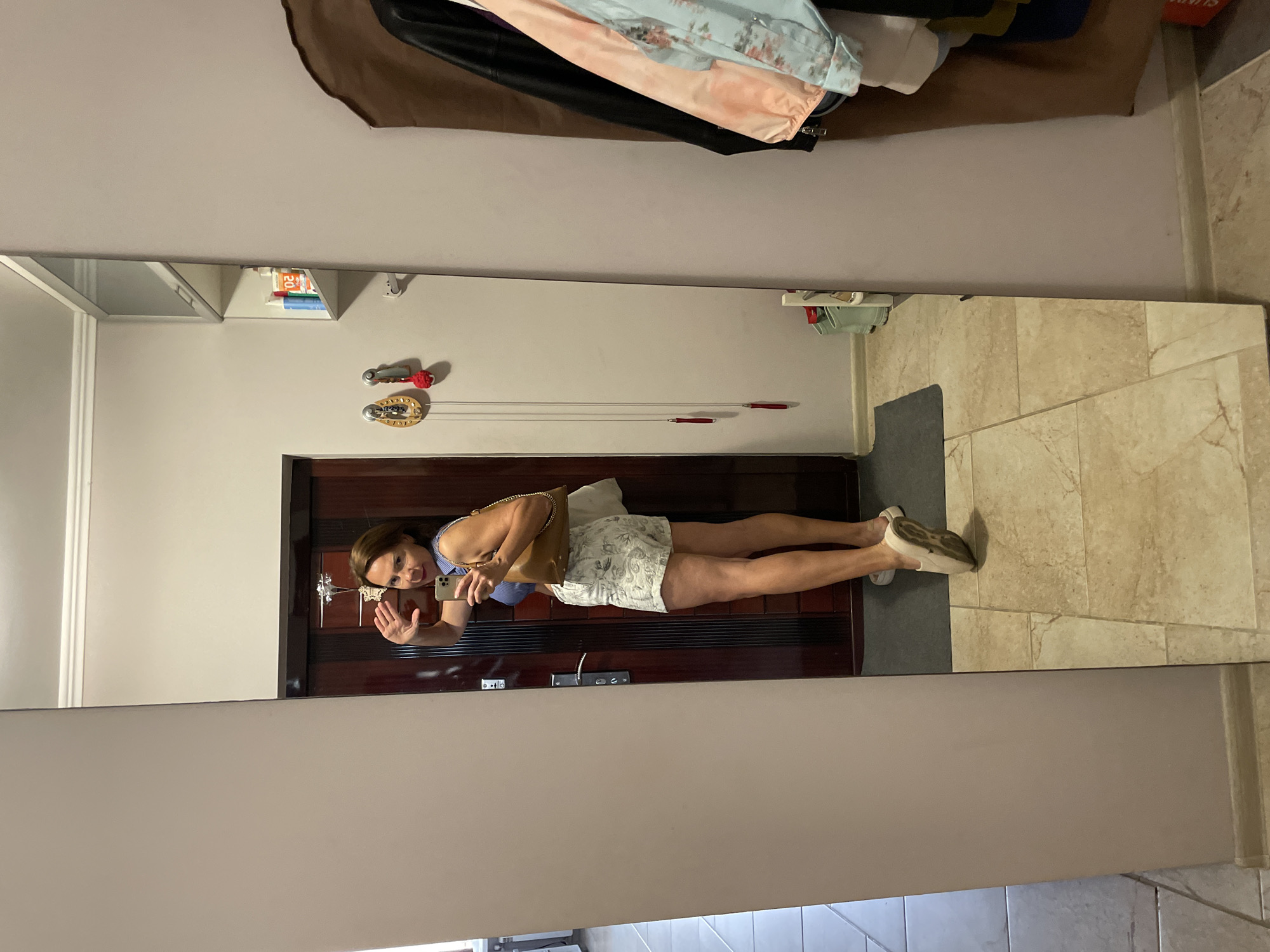 A woman stands in front of a full-length mirror in what appears to be an entryway or hallway, posing for a photo. She is wearing a blue and white striped top, white shorts with a light-colored pattern, and light-colored slip-on shoes. She is holding a brown handbag over her shoulder and her left hand is raised, possibly waving or adjusting her hair. The mirror reflects the woman, a dark wooden door behind her, a tiled floor, and a portion of a wall with some decorative items and shelving. On the right side of the image, a wardrobe or coat rack is visible with several jackets hanging on it, including a black jacket, a coral-pink jacket, and a light blue jacket with a floral pattern. The overall environment suggests a residential setting, possibly an apartment or house. There is no identifiable information within the image to confirm the location as Sochi, Russia.