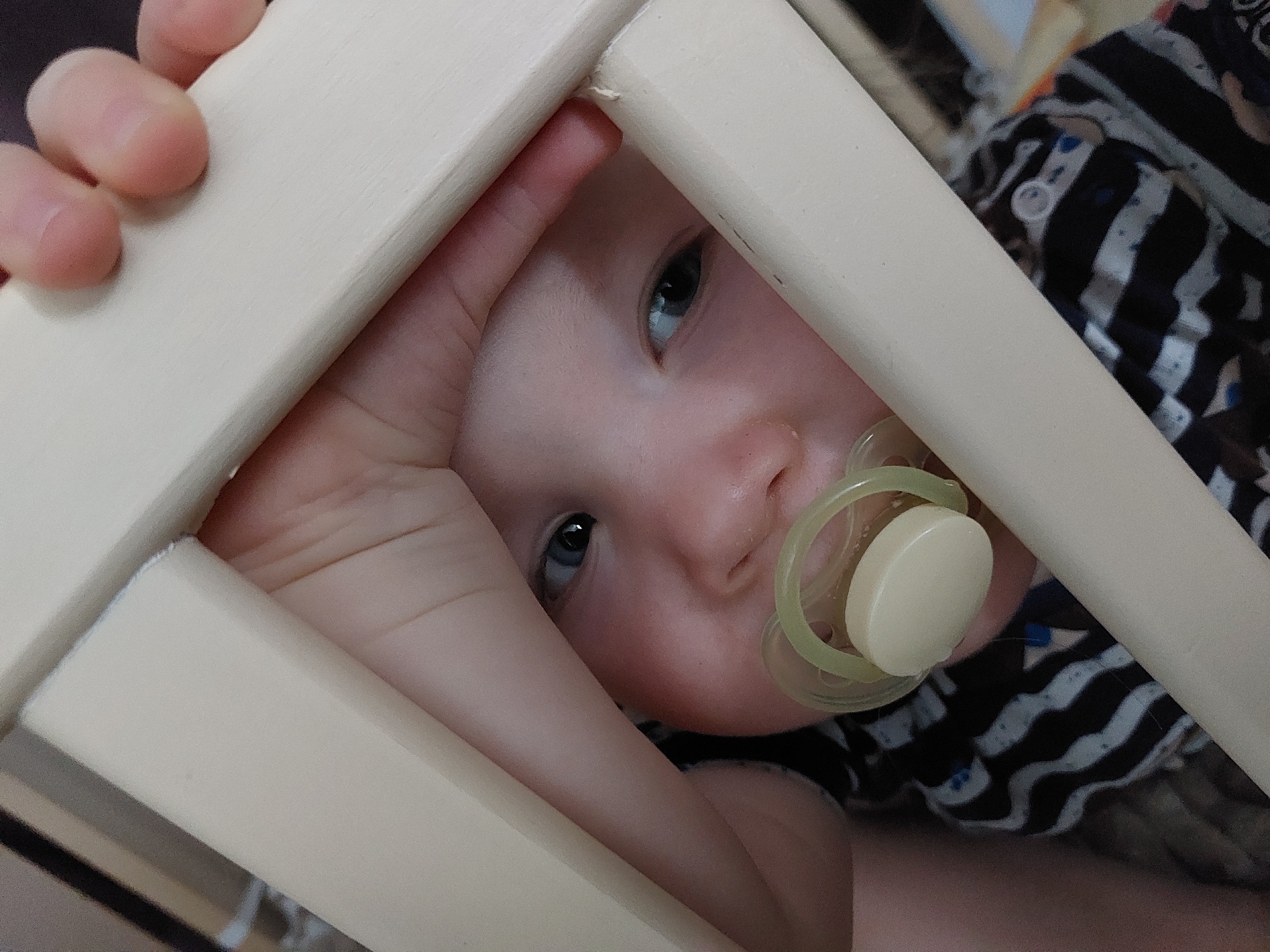 A baby with a pacifier in their mouth is peering through the bars of a cot. The baby's face is centered in the image, with their eyes looking directly at the camera. A hand grips one of the bars of the cot. The baby is lying down, with their body visible through the gaps in the cot bars. The background is blurry, suggesting a domestic setting. The location and city are unknown.