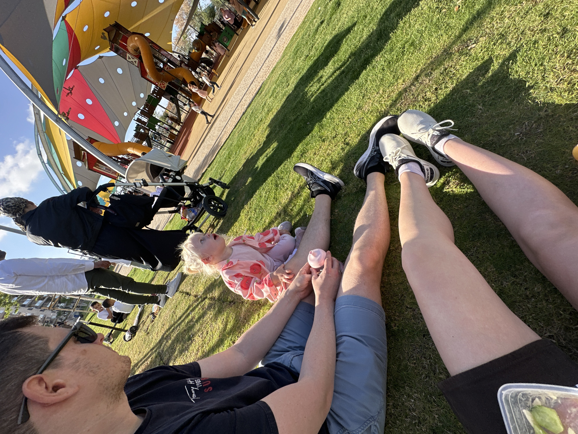 A photograph taken outdoors on a sunny day, likely in the city of Petah Tikva, Israel, depicts a family scene on a grassy area. A man in a black T-shirt and blue shorts sits with his legs extended, holding a pink bottle, and a young girl in a pink and white patterned outfit lies on his legs. Another person, possibly a woman, sits nearby with legs extended, wearing black shorts. Two pairs of sneakers are visible near the legs. Behind the family, people are standing. In the background, there is a stroller and a playground structure with colorful shade canopies.