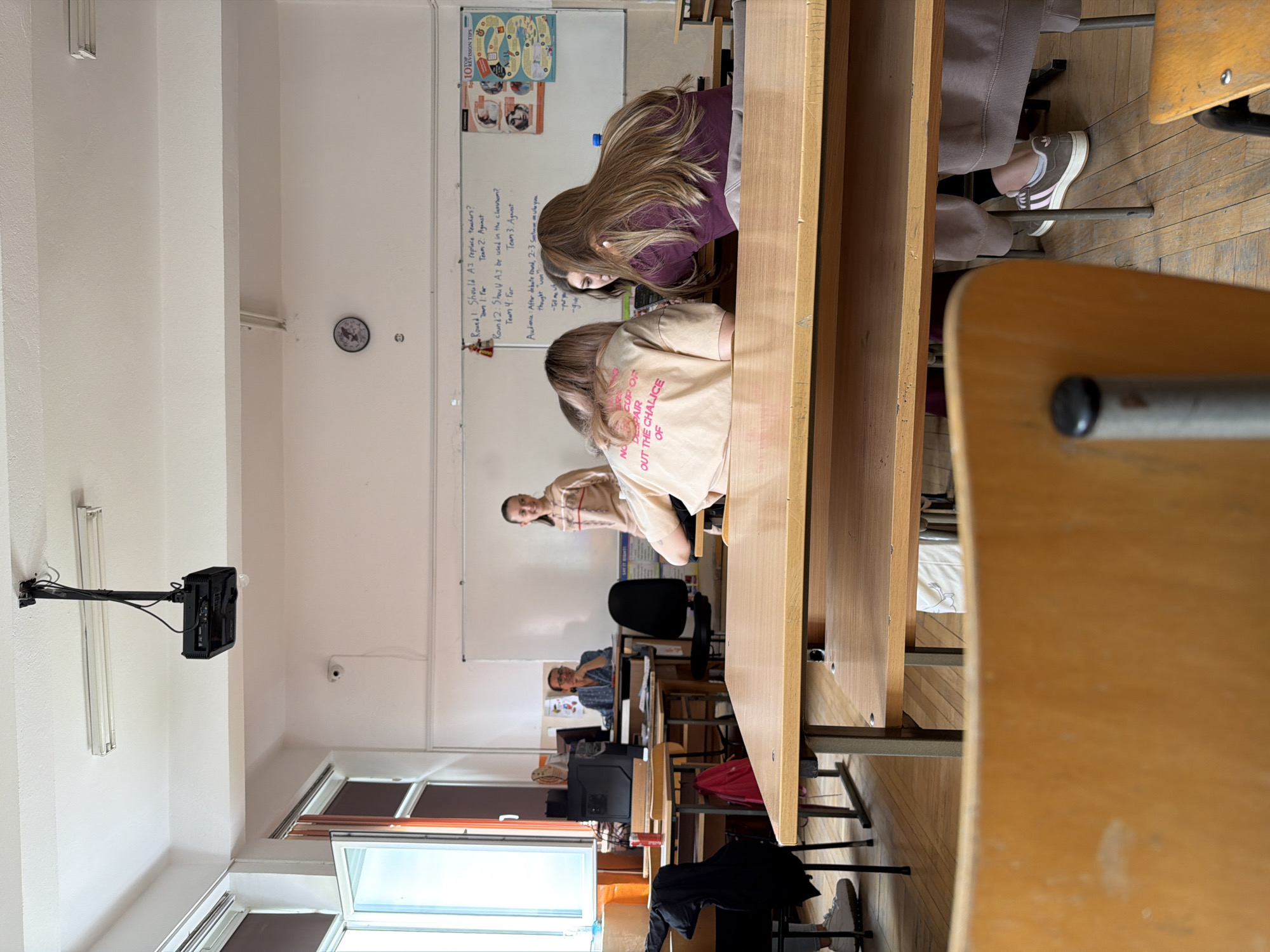 The image captures a classroom scene in Panagyurishte, Bulgaria. Several students are seated at wooden desks, with two students in the foreground closely examining something on their desk. A woman stands at the front of the class, seemingly instructing or observing the students. Another person is visible towards the back of the classroom, seated at a desk. A whiteboard at the front of the room contains written text and diagrams. The classroom is illuminated by overhead fluorescent lights. A clock is on the wall, and a window provides natural light. Desks and chairs are arranged in rows. Some personal items, like bags and clothing, are scattered around the floor.