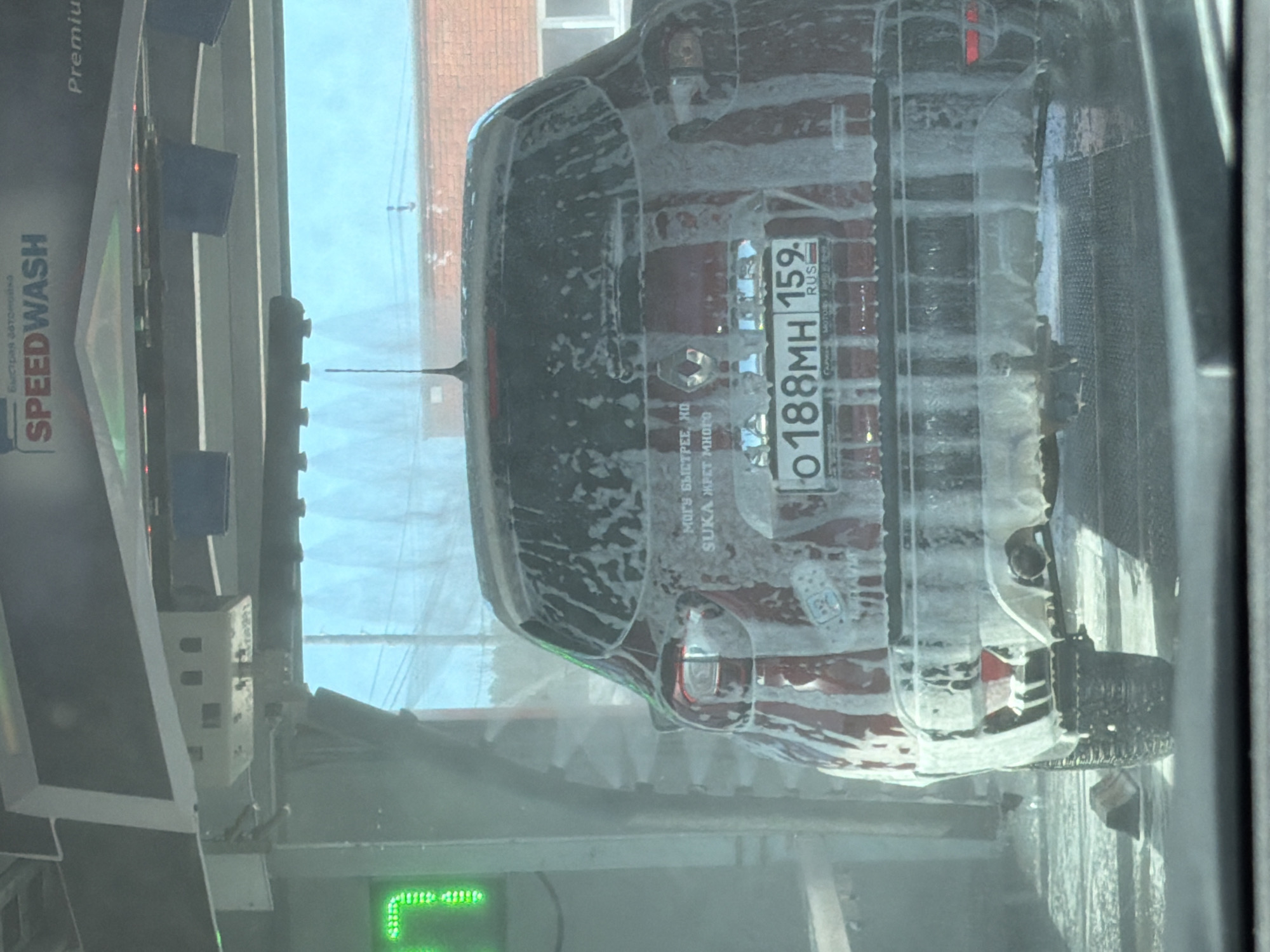 The image shows the rear of a dark-colored car going through a SPEEDWASH carwash. The car's license plate reads "O188MH 159 RUS." Text on the car reads: "МОГУ БЫСТРЕЕ, НО SUKA ЖРЕТ МНОГО" which translates to "I can go faster, but the bitch consumes a lot". The car has a Renault logo. The SPEEDWASH sign indicates a quick car wash service. The location, based on the license plate region code "159," is likely in the Perm Krai region of Russia, which includes the city of Chaikovsky.