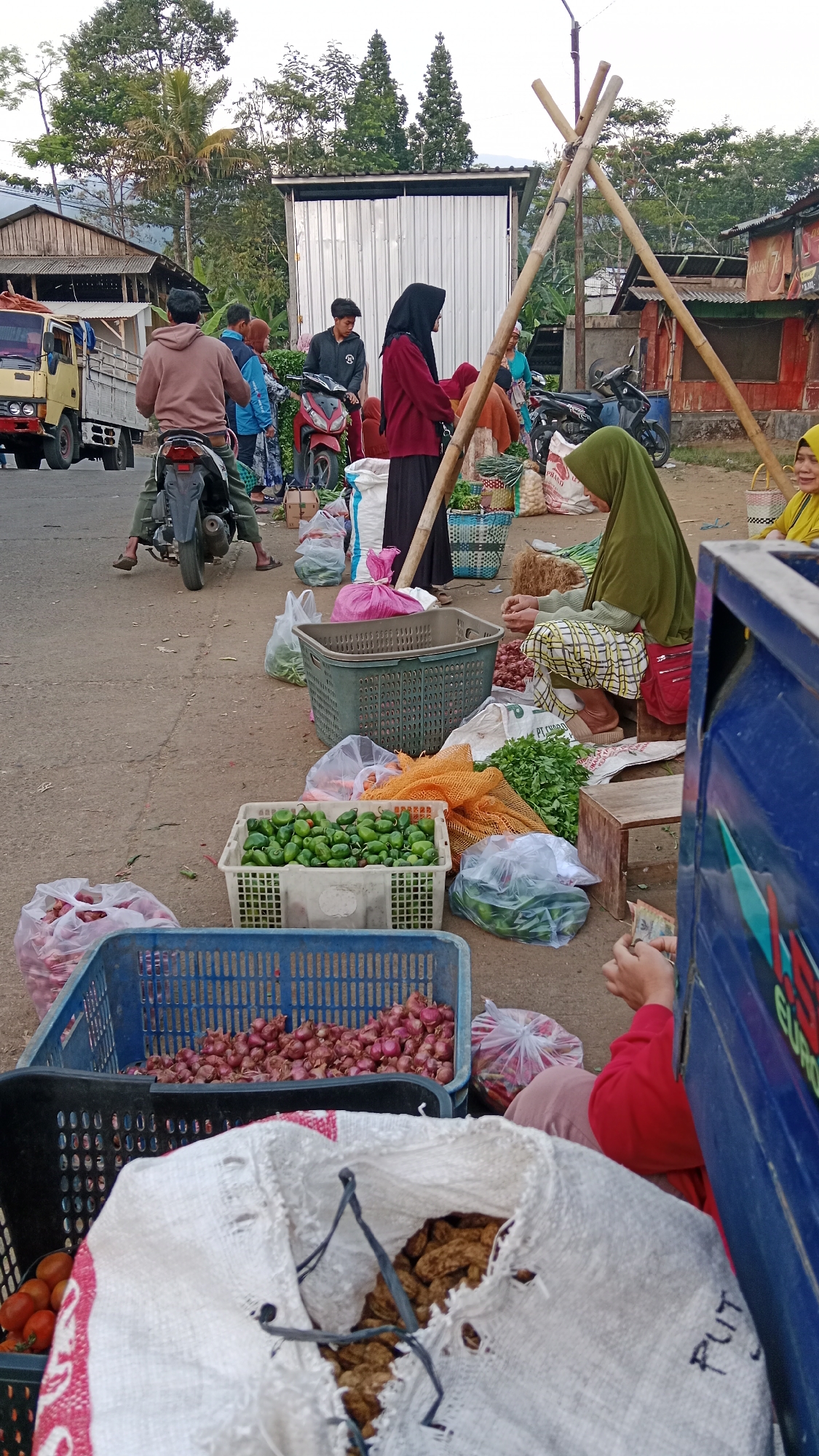 This is a street market scene in Klawen, Indonesia. A blue truck is parked on the right, with its side and part of its front visible. Several people are engaged in commerce, with baskets of produce laid out on the ground. In the foreground, a large white sack with red trim is open, revealing brown, dried goods. Next to it, a blue plastic basket contains red onions, and a black plastic basket holds more red onions. A grey plastic crate is filled with small green peppers. In the middle ground, a woman wearing a green hijab and patterned pants sits on a wooden stool, tending to the vegetables. She is handling money. Behind her, a dark-haired man in a black jacket stands near a red motorcycle. To his left, a woman in a maroon headscarf and black skirt stands, and further left, a man on a motorcycle wears a brown hooded sweatshirt. A yellow truck is visible on the far left of the image. In the background, buildings with corrugated metal roofs line a street, and lush green trees and vegetation suggest a rural or semi-rural setting. The ground is paved with asphalt.
