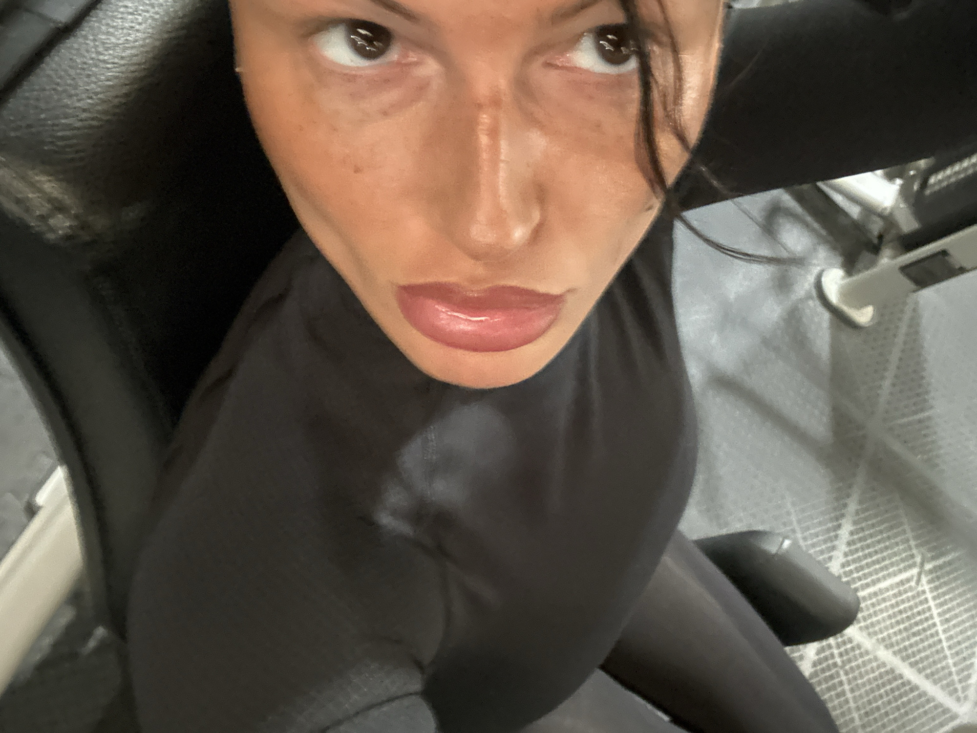A person is shown in a close-up, high-angle view. They are wearing a black, tight-fitting athletic shirt or bodysuit. The background appears to be a gym or fitness facility, with exercise equipment and a textured floor visible. The person is facing directly into the camera. No specific location within Tunbridge Wells is identifiable.