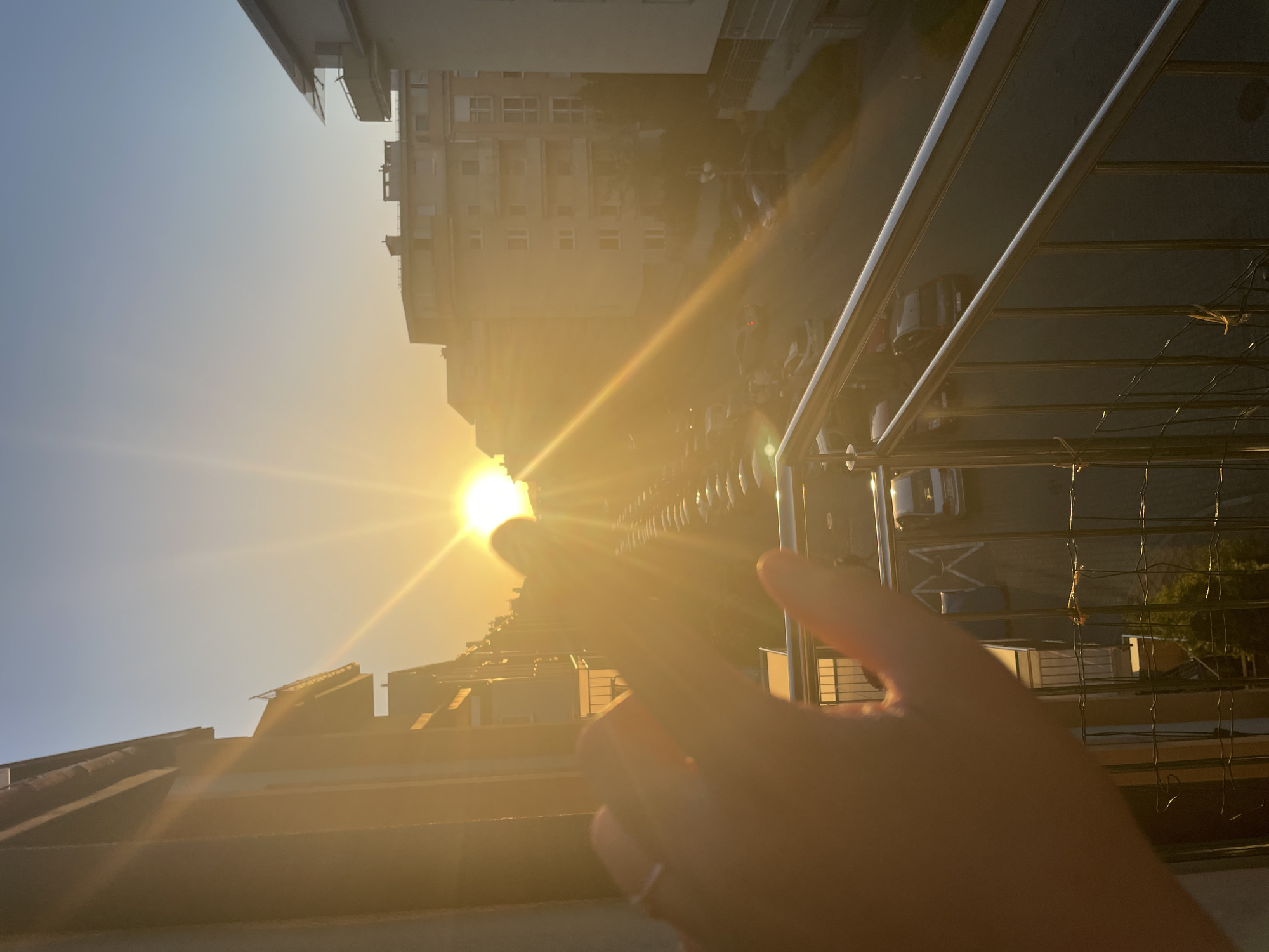 The image shows a hand pointing toward the sun. The backdrop is a cityscape with buildings and a street visible. The picture is taken from a high vantage point, possibly a balcony, as a railing is visible in the foreground. The lighting suggests it is either sunrise or sunset. The exact location in the city of Unknown is not possible to determine from the image.