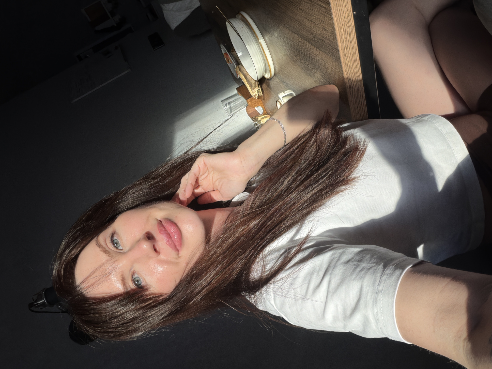 A woman is lying on a dark surface next to a table in what appears to be an indoor setting. The table holds a collection of items, including dishes, cutlery, a glass, and what looks like an incense burner. The woman has long, dark hair and blue eyes, and she is wearing a white t-shirt. Sunlight is casting harsh shadows on the scene. There is no indication that this image was taken in the city of Perm, Russia.