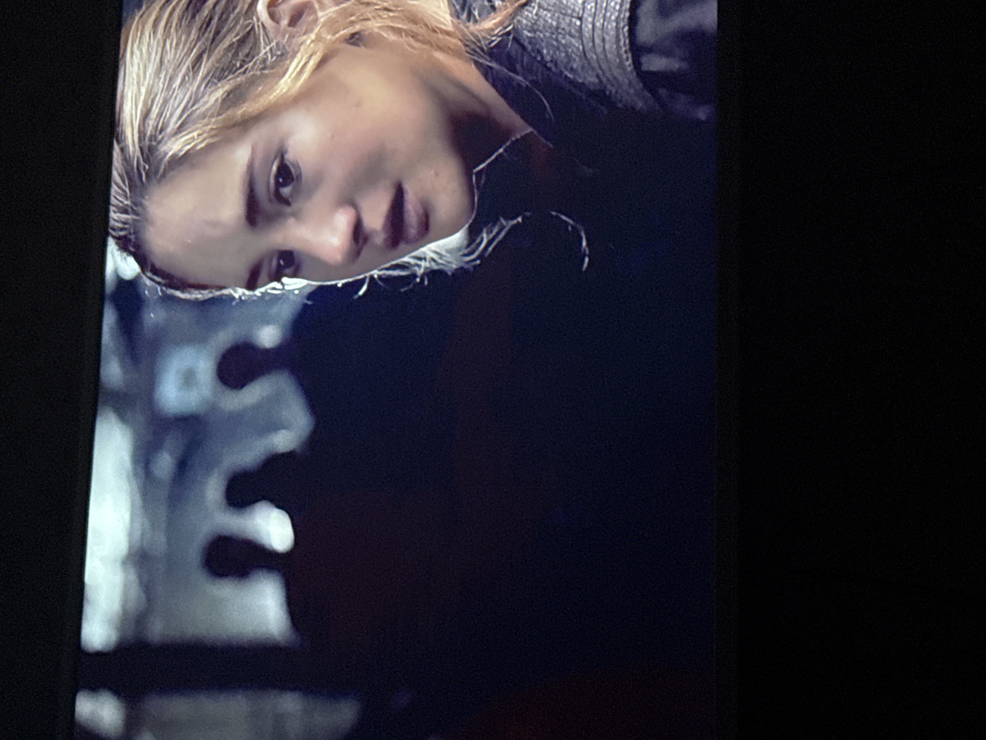 A low-angle close-up shot of a woman with wet, blonde hair and fair skin. She is wearing a dark, textured garment. Her expression is solemn and she is looking down. In the lower left of the frame, there is a blurred, abstract, greyish object. The background is dark and mostly out of focus. There is no location information.
