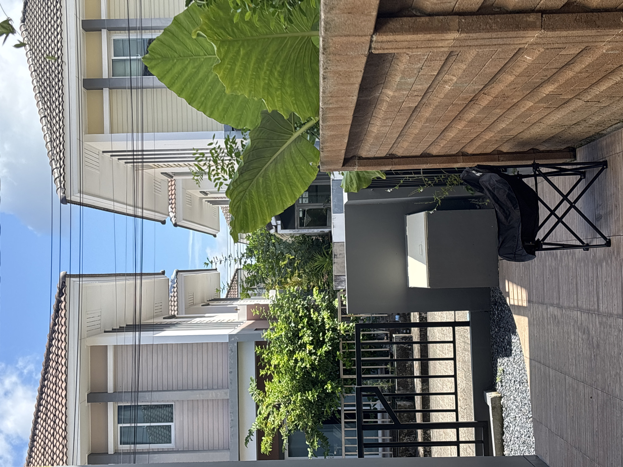 The image shows a residential scene in Ko Kaeo, Thailand. Two modern houses with beige and white siding and tiled roofs are visible, separated by a narrow gap. In the foreground, a black folding chair is positioned on a tiled patio. To the left of the chair is a grey rectangular bin with a white lid. A black metal fence with vertical bars runs along the edge of the patio. Beyond the fence, there is a patch of grey gravel. Two large, green elephant ear leaves are prominently displayed in the upper right of the image, obscuring part of the house on the right. The sky is blue with scattered clouds. Lush green trees and foliage are visible between and behind the houses. The scene suggests an outdoor living space or entrance to a residential property.