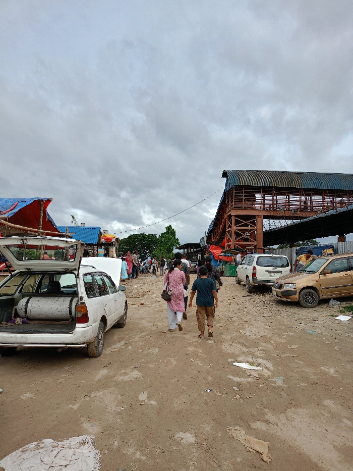 This image captures an outdoor scene on an unpaved, dirt thoroughfare in Sujabad, Bangladesh, under an overcast sky. The ground is uneven, displaying patches of dirt, gravel, and scattered debris.
On the left, a white station wagon is parked with its rear hatch fully open, revealing a large white cylindrical tank, likely for fuel. The vehicle's hood is also slightly ajar. Beyond the vehicle, temporary structures with blue and red tarpaulin coverings are visible, suggesting market stalls.
In the midground, a woman wearing a light pink top and white pants walks away from the viewer, accompanied by a male child or young man in a dark blue shirt and brown shorts. Both are walking on the dirt path. Numerous other individuals are visible further down the thoroughfare, either walking or standing, some in traditional attire.
On the right side of the frame, an elevated structure with a blue corrugated roof and reddish-brown metal framework extends into the background, resembling a pedestrian bridge or railway platform. Below this structure, a white minivan and a gold-colored sedan are parked on the dirt. A person in a yellow shirt is visible near the gold sedan. The background includes green trees and distant structures.