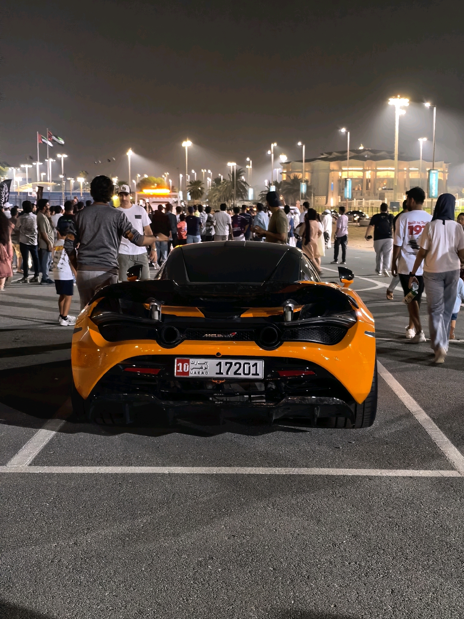 An eye-level, medium shot captures the rear of an orange and black McLaren sports car parked in a paved lot at night. The car's license plate, reading "10 UAE 17201", is clearly visible. The background is populated with a crowd of people, some walking and others standing. Beyond the crowd, illuminated buildings and streetlights suggest an urban or entertainment area. Two flags, one green and white and the other red, white, and black, are visible in the distance, hinting at the location being the United Arab Emirates. The overall scene suggests an event or gathering taking place outdoors after dark.