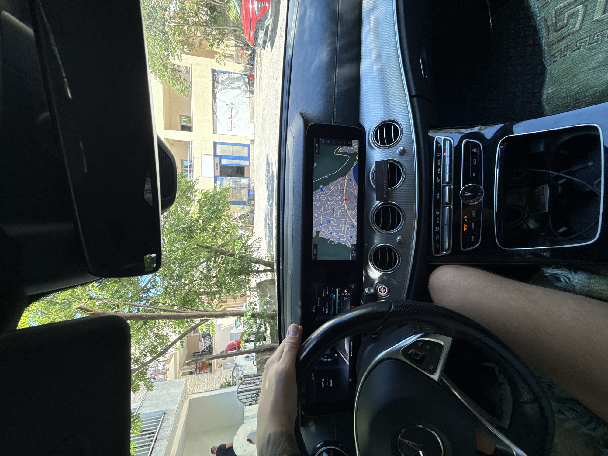 The image, taken from the interior of a Mercedes-Benz E-Class, shows a driver's hand on the steering wheel. The car's dashboard features a large infotainment screen displaying a navigation map of Budva, Montenegro, with the car's current position marked. The view outside the windshield reveals buildings and trees. To the left, two men are visible near a building, and a red car is parked on the right. The overall scene depicts a moment inside a vehicle on a street in Budva.