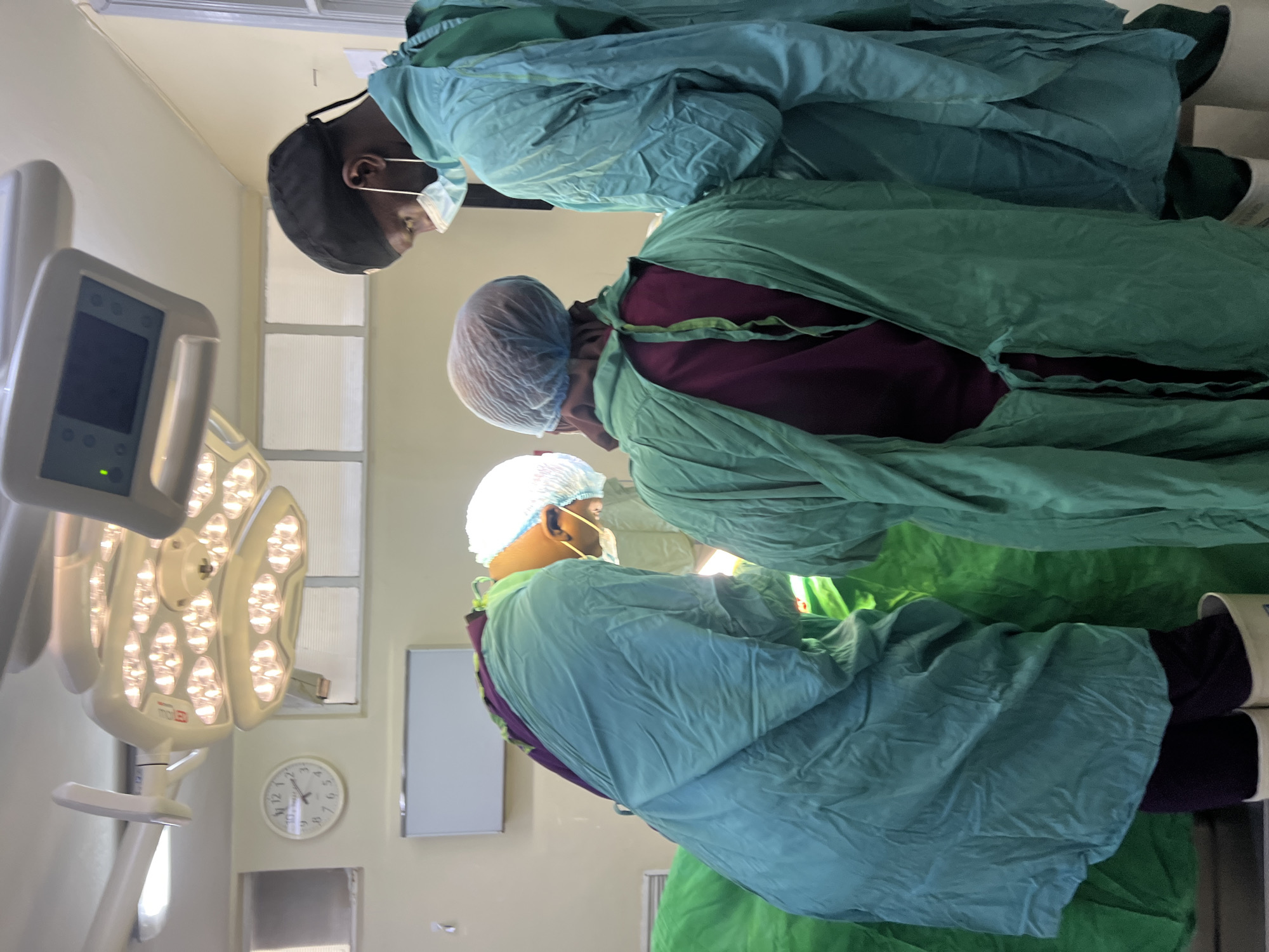 The image shows three individuals in an operating room setting, likely in a medical facility. All three are wearing surgical attire, including surgical gowns, caps, and masks. They appear to be focused on a surgical procedure in progress. The room is equipped with a surgical lamp, a monitor, and a clock on the wall. The presence of a person of African descent suggests the location might be in Nairobi, Kenya.