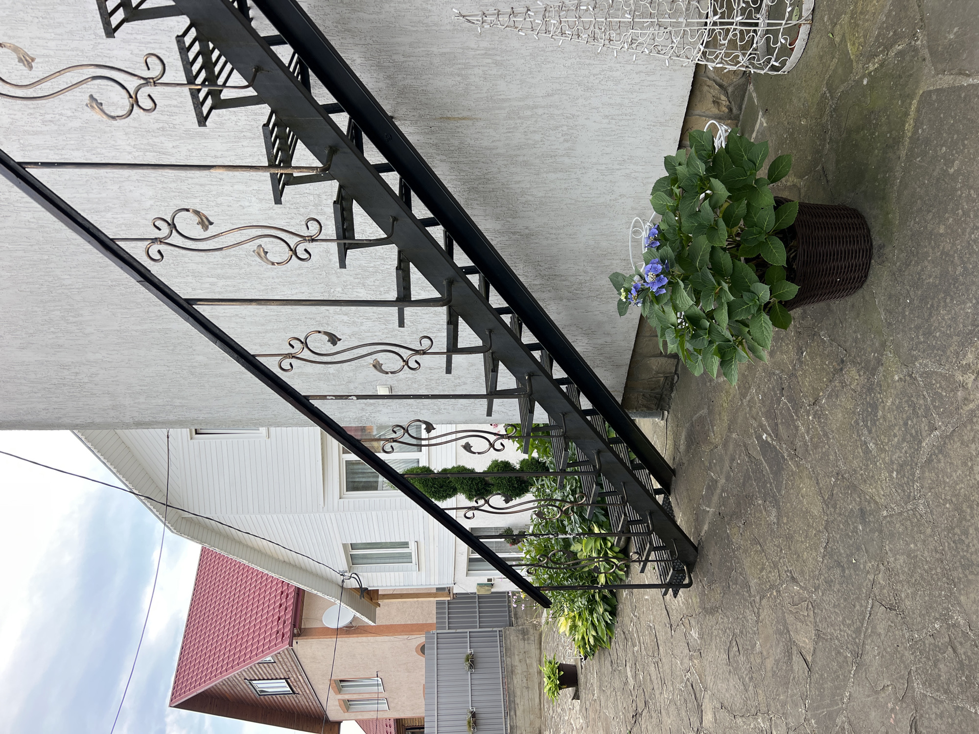 The image depicts an outdoor scene in Yaremche, Ukraine, featuring a section of a building with an exterior metal staircase. The staircase is black with ornate metal railings. In the foreground, a potted plant with blue flowers sits on a stone patio. To the left, a grey metal fence encloses a small garden area with lush green plants. In the background, two houses are visible; one is white with a red roof, and the other is beige with a red roof. The sky is overcast. The overall impression is of a residential outdoor space.