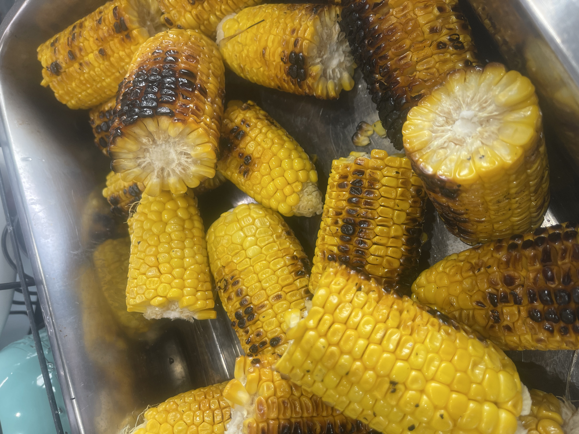 The image shows a close-up, overhead view of several ears of corn that have been cut into segments and appear to be grilled. The corn is bright yellow with some sections showing charring from the grilling process. The corn is arranged in a metal tray, which is likely stainless steel. The background is slightly out of focus but shows what appears to be a kitchen or food preparation area with some metallic surfaces and a hint of a teal-colored object. There are no people visible in the image. The specific location in the city of Karlovo, Bulgaria cannot be determined from the provided image.
