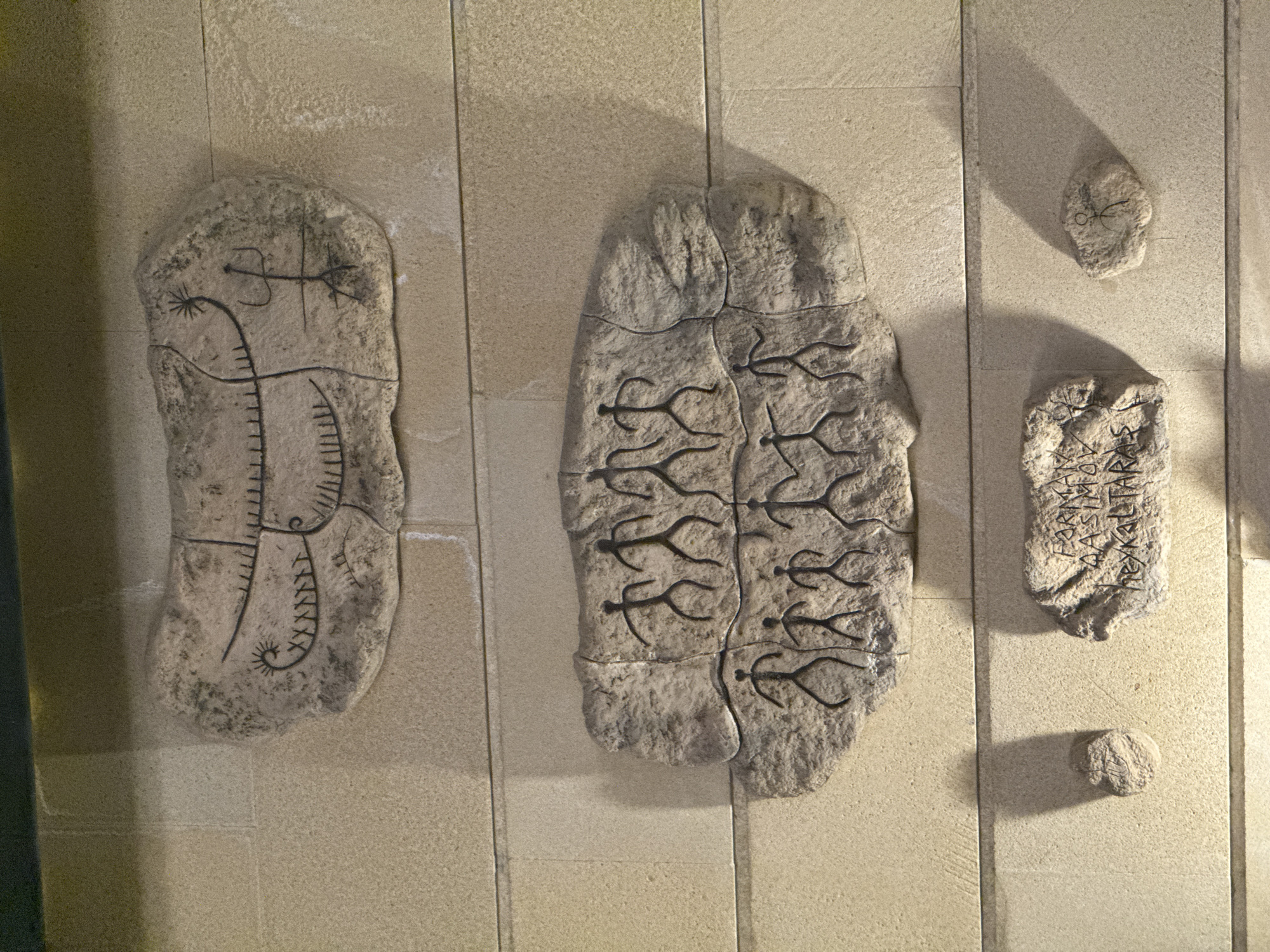 The image displays a series of stone reliefs mounted on a textured, light-colored wall, reminiscent of sandstone blocks. The uppermost relief depicts a stylized boat with a sun-like symbol and a human figure. Below this, two larger reliefs showcase rows of stick-figure humans, some appearing to be in motion or engaged in an activity. The bottom section of the image features two smaller stones. One bears faint etched markings that appear to be text, possibly in Greek, and the other has a single, small stick-figure drawing.
These reliefs are located within the **Gobustan National Park in Baku, Azerbaijan**. The park is renowned for its extensive collection of rock carvings, known as petroglyphs, which date back thousands of years and offer a glimpse into the prehistoric life and culture of the region. The wall and the way the stones are displayed suggest an exhibition or museum-like setting within the park, designed to present these ancient artifacts.