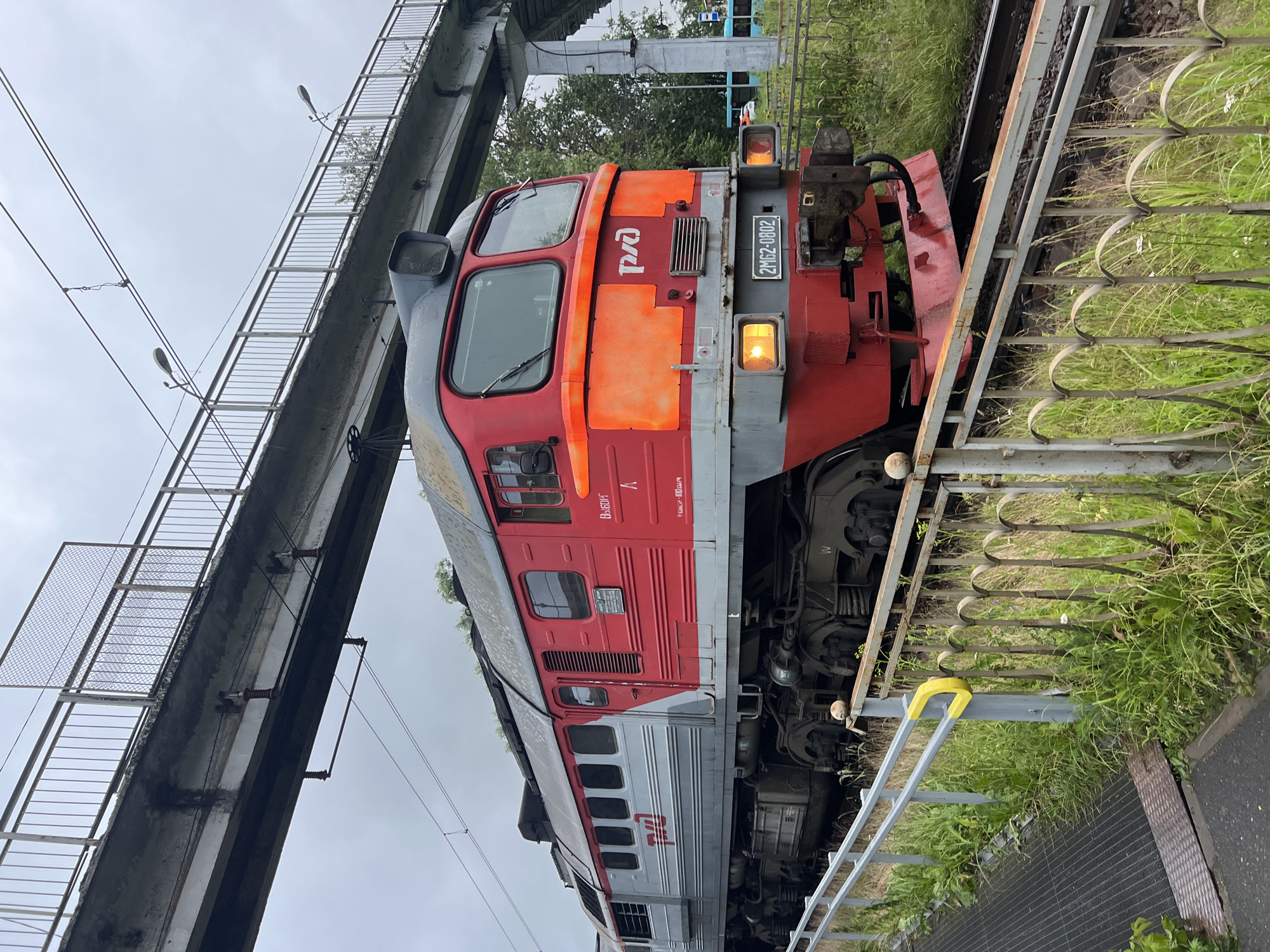 An outdoor, low-angle shot shows a red and gray diesel locomotive positioned on train tracks in Alexandrovskaya, Russia. The locomotive, bearing the Russian Railways logo "РЖД" and the identification number "2М62-0802", is partially obscured by a metal railing in the foreground, which is surrounded by lush green grass. A pedestrian bridge with a metal fence and overhead lights extends across the upper portion of the frame, over the train tracks. The sky is overcast.