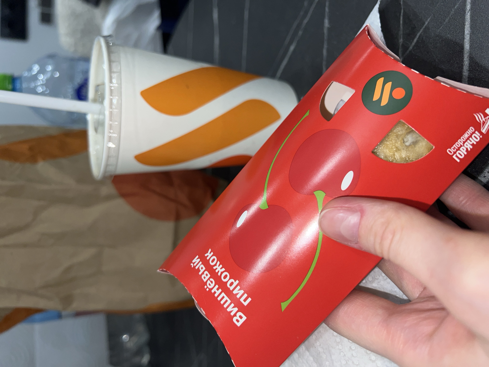A photograph shows a McDonald's cherry pie box held in a person's hand alongside a partially visible beverage cup. The pie box is red, displaying a stylized image of cherries and text in Russian. The text translates to "Cherry Pie" and includes a warning "Careful! Hot!". The McDonald's logo is also present on the box. The beverage cup is white with orange stripes. A clear plastic lid and a white straw are visible on the cup. A partially visible water bottle is present behind the cup. The scene is set indoors, presumably on a table, and based on the products and language, it is likely a McDonald's location in Khimki, Russia.