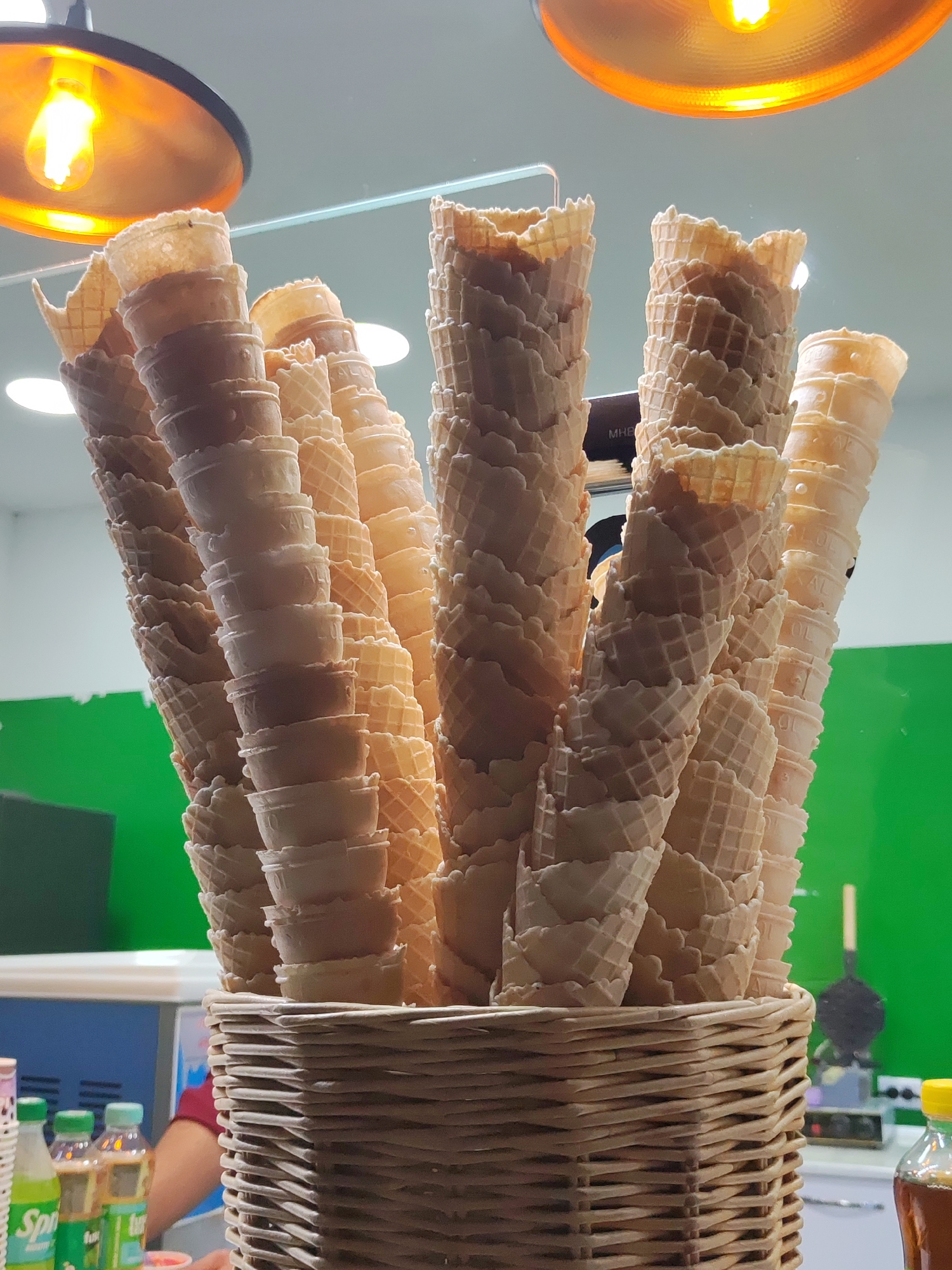 A close-up shot captures a wicker basket overflowing with stacked waffle ice cream cones. The cones, in various shades of light brown and beige, are neatly arranged in vertical stacks, with some wider at the rim and tapering towards the bottom. The textured pattern of the waffle cones is clearly visible. Behind the cones, a bright green wall serves as a backdrop, and a hint of a metal waffle maker can be seen to the right, suggesting a food service environment. Two bottles of a beverage, one labeled "Sprite," are partially visible in the lower left corner, along with a portion of a person's arm and shoulder. The scene is illuminated by warm, yellow lighting from above, likely from pendant lamps. The setting is a food stall or cafe, possibly in Tashkent, Uzbekistan.