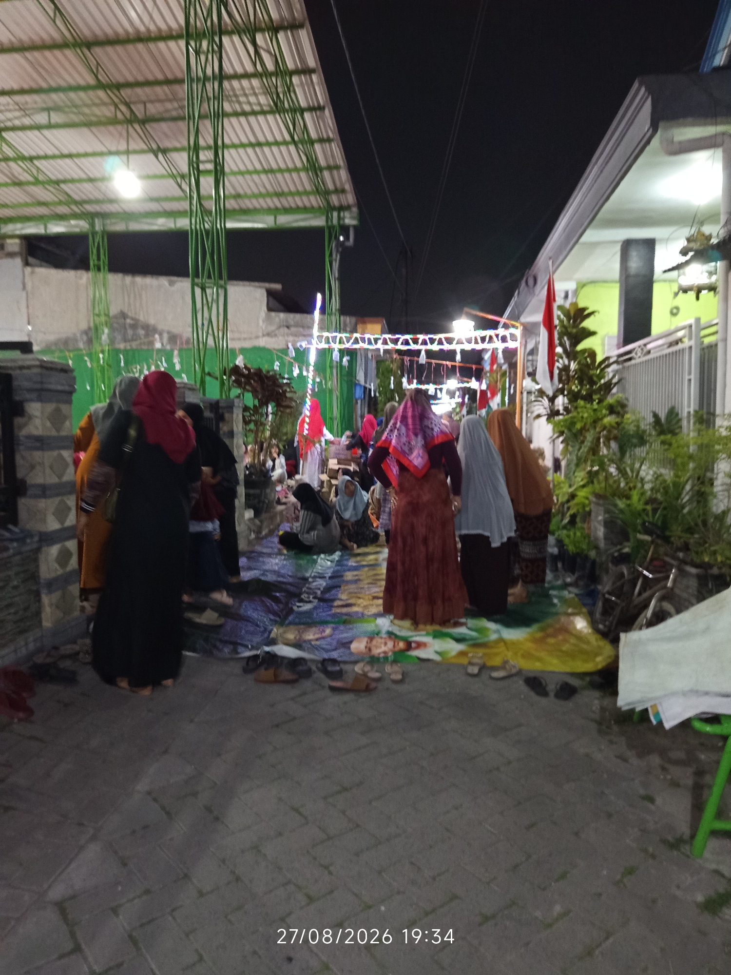 The image depicts an outdoor gathering at night, likely in Canggu, Indonesia, given the timestamp and context. The scene is lit by artificial lighting, casting a yellowish glow. Several people, predominantly women wearing hijabs and modest clothing, are present. Some are seated on the ground on mats, while others are standing. One individual in the foreground wears a red hijab and a dark garment, while another to their left wears an orange garment and a grey hijab. In the background, more people are visible, some appearing to be children.
The environment features a covered outdoor area with a corrugated metal roof supported by green metal pillars. There are also decorative lights strung overhead. A white fence is visible on the right side of the image, bordering a house with green walls. Potted plants, including a large leafy plant, are situated near the fence. On the ground, there are various pairs of footwear, including sandals and flip-flops. The ground is paved with interlocking blocks. A large white tarp or sheet is present in the lower right corner. The overall impression is of a community event or religious gathering taking place after dark.