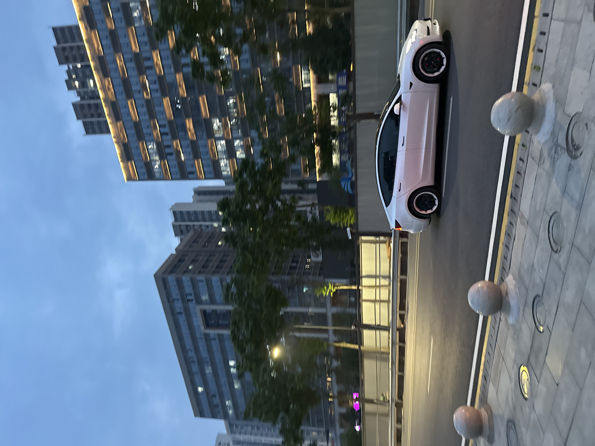 A white Tesla sedan drives on a wet road in the Chongqing High-tech Zone, China, under a cloudy twilight sky. The car is in motion, with its headlights off and windows tinted. The road is paved and appears to be a multi-lane street with yellow and white lines marking the lanes. In the background, modern buildings with many windows are illuminated, suggesting it is evening. Trees line the street, and there are decorative spherical bollards along the sidewalk. The overall impression is of a contemporary urban environment at dusk.