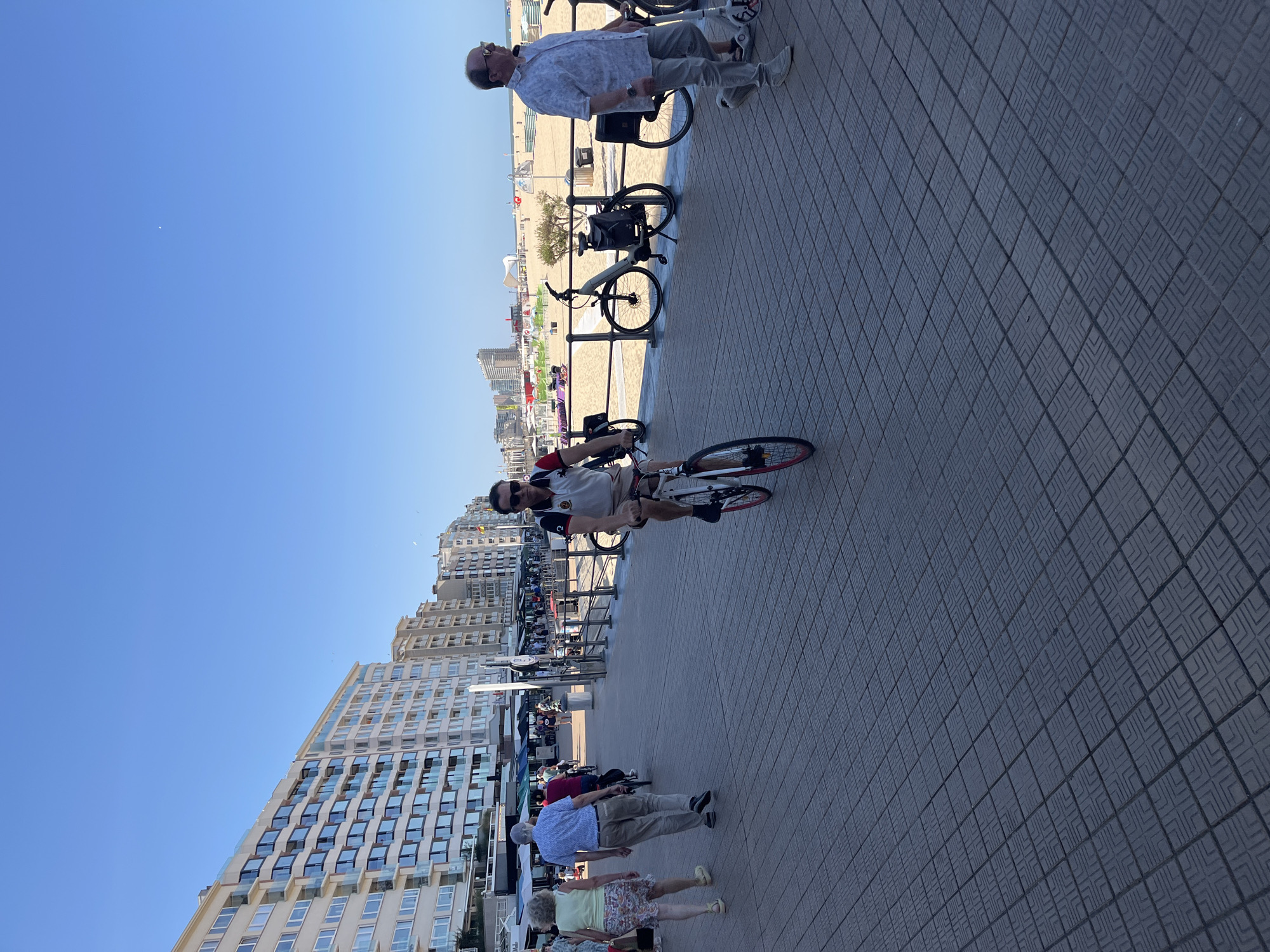 The image depicts a sunny day on the Ostend, Belgium waterfront promenade. A man in a white polo shirt with red and black trim is cycling towards the viewer on a white bicycle with red rims. He wears sunglasses and has short, dark hair. To his left, a couple walks away from the camera; the woman wears a light-colored top and a patterned skirt, while the man wears a blue patterned shirt and khaki pants. Further back, other pedestrians and cyclists are visible on the promenade, with buildings lining the left side of the image and the beach and ocean on the right. Several bicycles are parked along a railing separating the promenade from the beach. The promenade is paved with dark, square tiles.