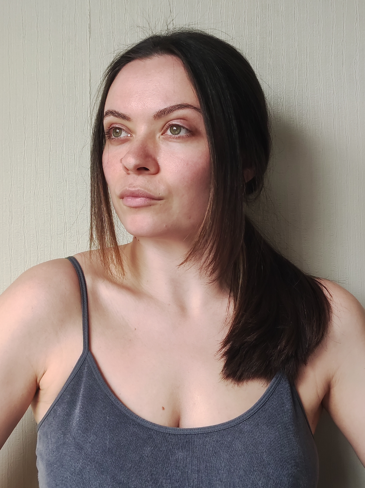 A young woman with dark brown hair, pulled back in a low ponytail, is wearing a gray ribbed tank top. She is looking to the left of the frame. The background is a light beige wall with a subtle vertical texture. The location is Unknown, Unknown.