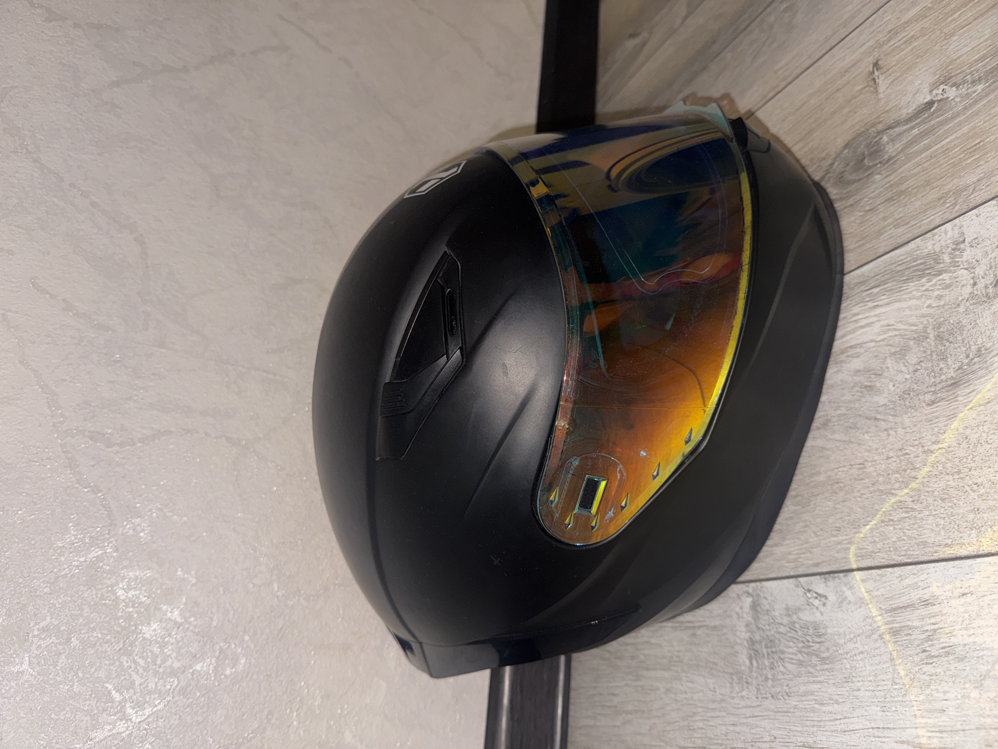 A black motorcycle helmet with a reflective iridescent visor rests on a light-colored wooden floor, leaning against a textured off-white wall. The helmet is positioned at an angle, with its front facing towards the right of the frame. The visor displays a rainbow of colors reflecting the surrounding environment. There are subtle scuff marks visible on the helmet's surface. The background wall has an uneven, textured finish, possibly wallpaper or painted plaster, with some faint darker lines and speckles. The wooden floorboards are laid horizontally and exhibit a light grain pattern. The overall lighting appears to be artificial and somewhat diffused. No people or actions are depicted in the image. The location is not discernible from the image itself; Bishkek City, Kyrgyzstan cannot be determined.