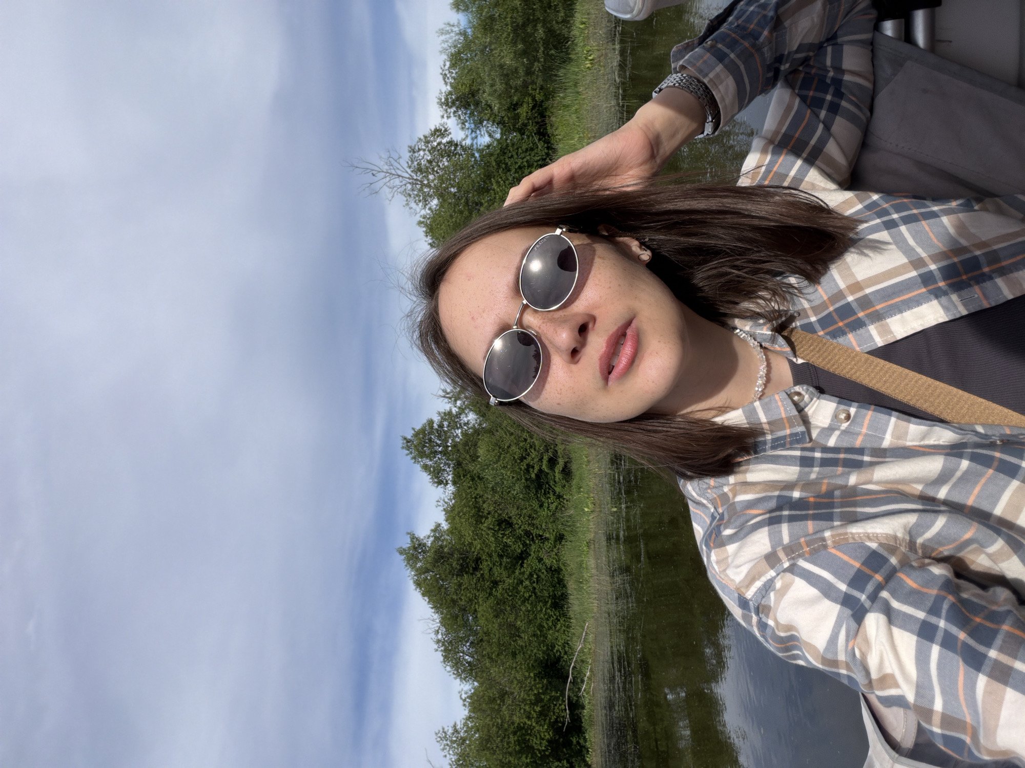 A young woman with brown hair and freckles is taking a selfie outdoors. She is wearing oval sunglasses, a plaid shirt, and a necklace. She has her hand on her head. The background shows a body of water, trees, and a cloudy sky. The scene appears to be in a natural environment.