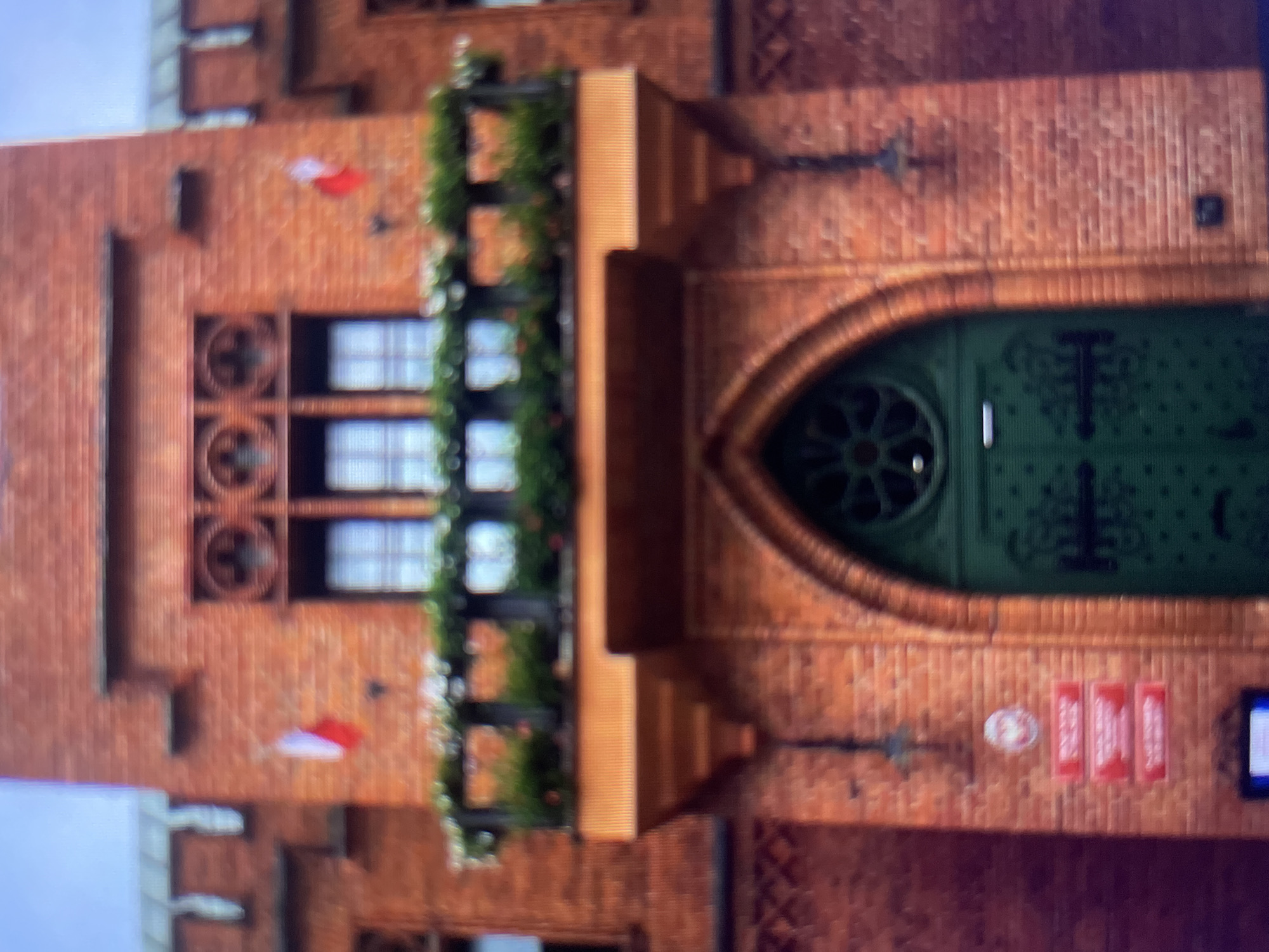 The image shows the exterior of a brick building with Gothic architectural elements. Two Polish flags are visible on the facade, one on each side of a balcony adorned with potted plants. Below the balcony, a large, ornate green door is set within an arched entryway. Several red signs with white text are mounted on the brickwork to the left of the door. The overall impression is of a historical or governmental building, possibly a town hall or a court. The architecture, particularly the crenellations and window designs, suggests a European setting. The presence of Polish flags indicates the location is in Poland. The quality of the image is low, with significant blur and pixelation, making precise identification of details difficult.