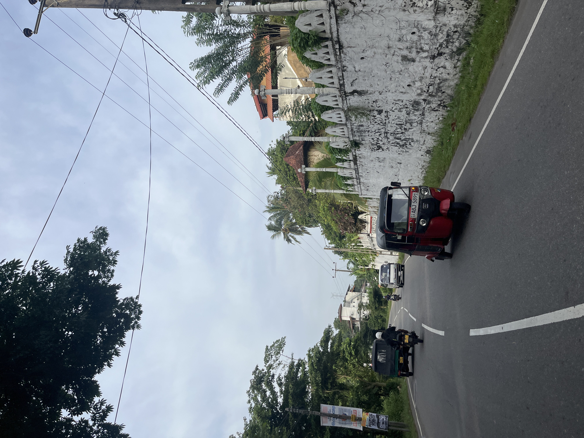 In Walahanduwa, Sri Lanka, a roadside scene unfolds under an overcast sky. A paved road, marked with a white dashed line, curves through a lush green environment. On the right side of the road, a textured white wall with decorative triangular cutouts runs alongside, topped with a railing. Behind the wall, a multi-story building with a red-tiled roof is visible amidst dense foliage.
Several vehicles navigate the road. A red three-wheeled auto-rickshaw, displaying the license plate "SB ABJ 5891," occupies the foreground on the right. Further down the road, a white van is in motion. A person on a motorcycle, wearing a white helmet, travels ahead of the van. On the left side of the road, a dark green three-wheeled auto-rickshaw is also moving. Alongside the road on the left, a tall pole supports multiple power lines that crisscross the sky. A billboard with text in a local script and English is affixed to a pole on the far left. Trees and vegetation line both sides of the road, with palm trees adding to the tropical atmosphere. The overall scene suggests a typical day on a road in this Sri Lankan locality.
