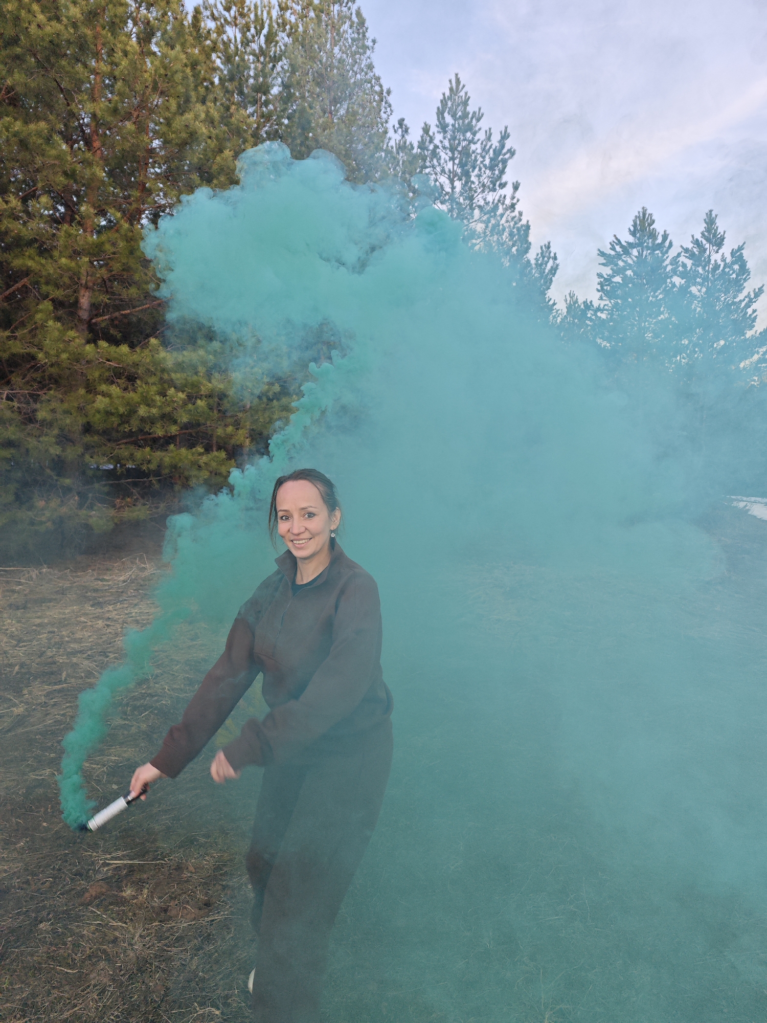 A woman is captured outdoors, actively releasing a large, vibrant teal-green cloud of smoke from a hand-held smoke grenade. She stands in a natural setting, with dense pine trees forming a backdrop behind her and dry, patchy ground cover underfoot. The sky is overcast and pale, providing soft, diffused light across the scene, hinting at either an overcast day or late afternoon.
The woman is wearing a dark, possibly brown or black, long-sleeved top and trousers. She holds the silver and white smoke grenade in her right hand, extended downwards and to her side, directing the substantial plume of smoke that billows around her, dominating the right half of the image and trailing behind her. She has a broad, joyful smile on her face, conveying a sense of pleasure or excitement. Her pose suggests she is in motion, possibly turning or walking as the smoke disperses. No discernible text is visible on the smoke grenade or elsewhere in the scene. The event takes place in Dubovaya, Russia.