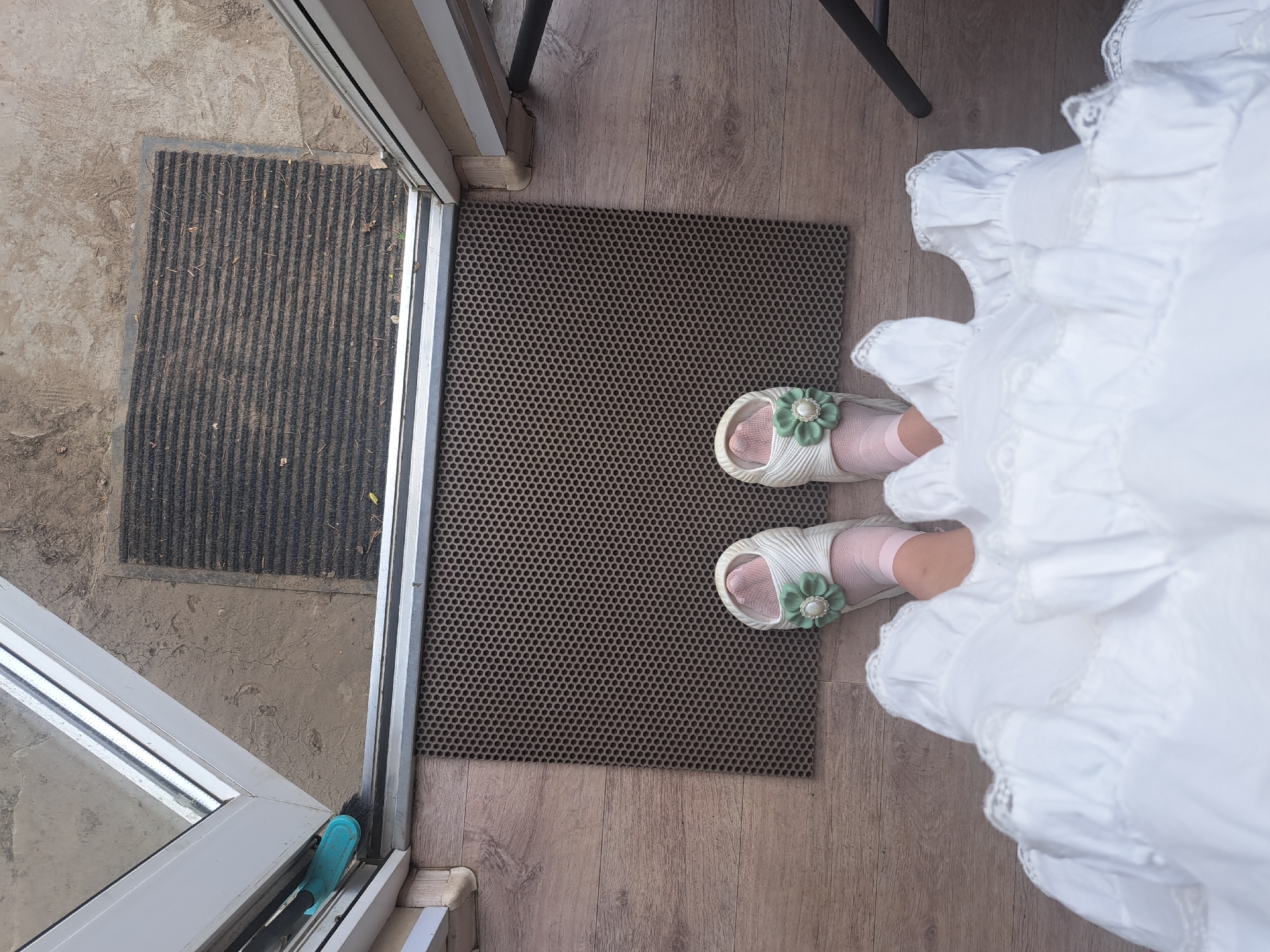 A top-down shot depicts a person standing on a brown, patterned doormat inside a building in Leningradskaya, Russia. The person is wearing white ruffled clothing and pink socks with white slippers adorned with green flower decorations. Part of a sliding glass door with a black doormat outside is visible to the left. A chair leg can be seen in the background. The floor is made of wood panels.