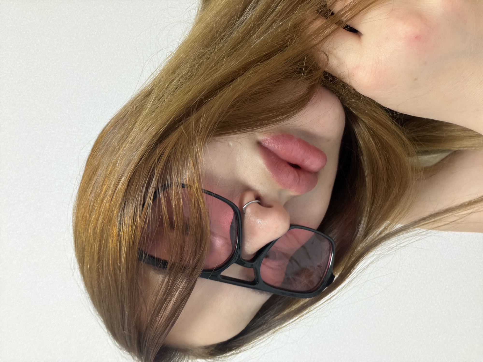 The image presents a close-up, angled shot of a person's head, tilted in an unusual orientation, possibly lying down or captured from above. The subject has long, light brown or auburn hair, which is somewhat disheveled and flows across parts of their face and the background.
Key facial features visible include their full, pink lips, which are slightly parted. A prominent detail is a silver hoop piercing in the right nostril. The eyes are obscured by both the hair and a pair of black-framed, square-shaped sunglasses with pink-tinted lenses, which are resting low on the nose. A portion of the person's fair-skinned hand or arm is visible in the upper right section of the frame, touching their hair.
The background is a plain, bright white or off-white, providing no specific location cues. There are no discernible activities taking place beyond the pose itself. The image appears to be an artistic or casual portrait, focusing on the interplay of hair, facial features, and accessories. No text is visible in the image.