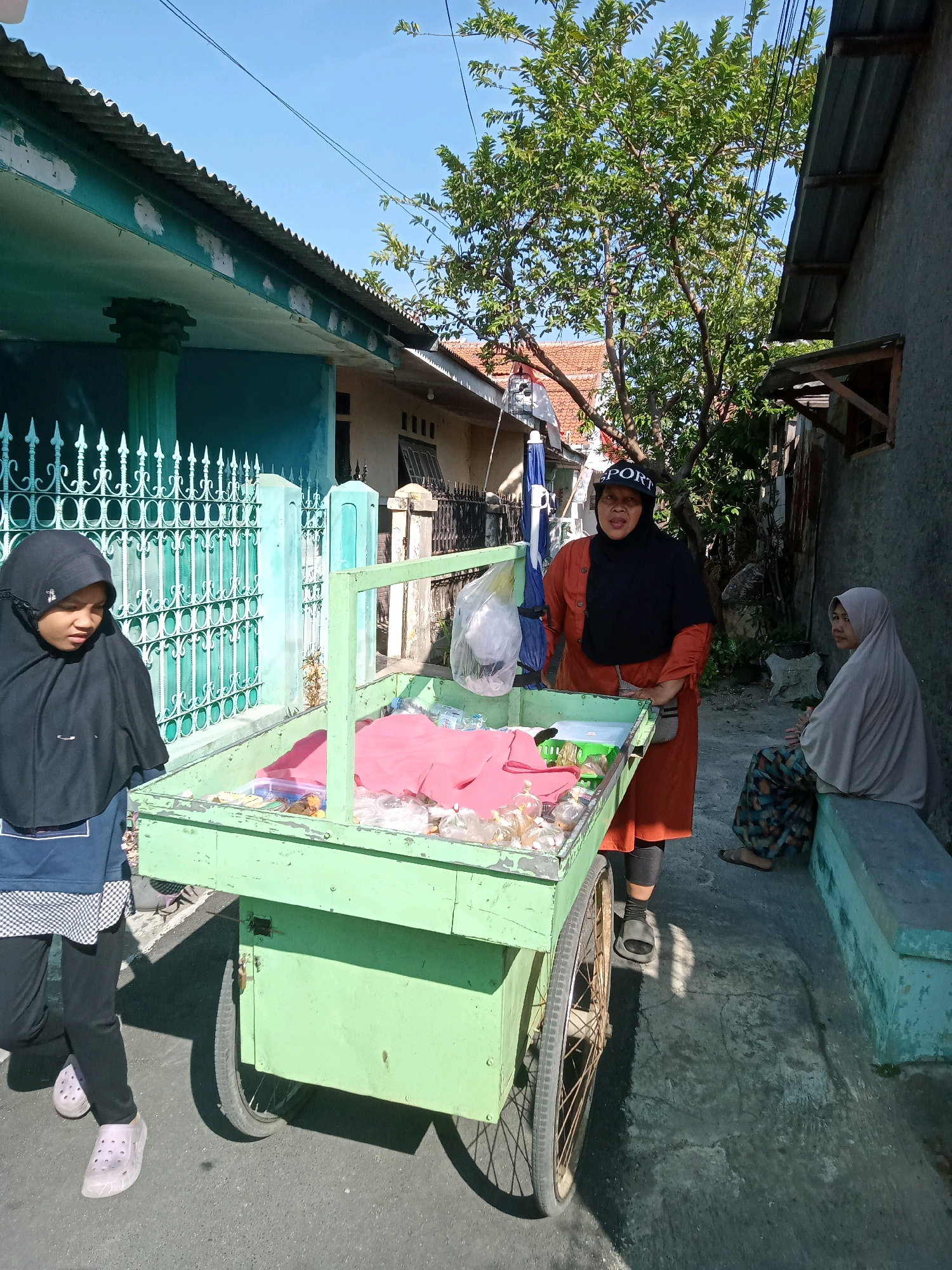 In Cirebon, Indonesia, under a bright, clear sky, a street scene unfolds. A woman in an orange tunic and black hijab, wearing a "SPORT" cap, stands beside a green, two-wheeled cart laden with goods. The cart's contents include pink fabric, bottles of water, and small containers of what appear to be snacks or trinkets. To the left, a young girl in a black hijab and blue tunic observes the cart. To the right, a woman in a beige hijab and patterned skirt sits on a low, blue-green concrete bench, observing the scene. The environment is characterized by the rustic architecture of residential buildings with corrugated metal roofs, a green metal fence, and a tree with lush foliage casting dappled shadows on the concrete ground. The image captures a moment of everyday commerce on a sunlit Indonesian street.