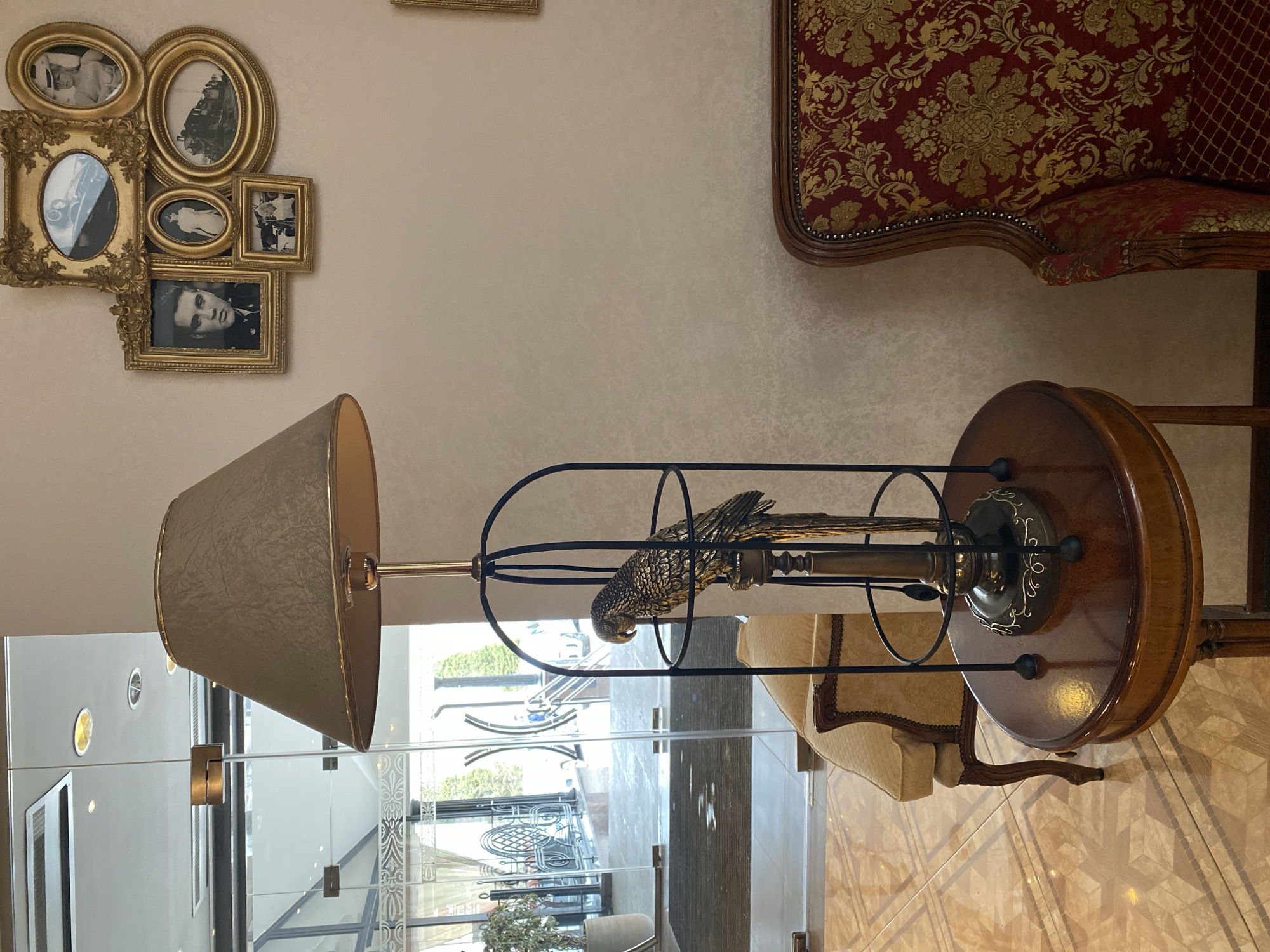 The image depicts an interior scene, presumably a high-end establishment.
Foreground: A distinctive lamp with a tan shade stands prominently. Its design features a metal bird within a cage-like structure attached to the lamp stand, all situated on a round wooden table. Behind the lamp, a tan upholstered chair is partially visible. The flooring has a geometric tile pattern.
Background: The view extends through what appears to be a glass wall or door into an outdoor area. In this outdoor space, decorative elements are visible, including green plants and metal railings. Further back, hints of the urban environment are discernible.
Upper portion: On the wall, there's a collection of ornate gold frames, containing black and white photographs. To the right, a section of a red upholstered chair with gold detailing is seen.
Overall: The environment appears to be luxurious and well-maintained. The presence of the glass wall/door suggests a transition between an indoor and outdoor space, likely within a building in an urban setting. The lamp serves as a focal point, highlighting the unique design elements of the interior.