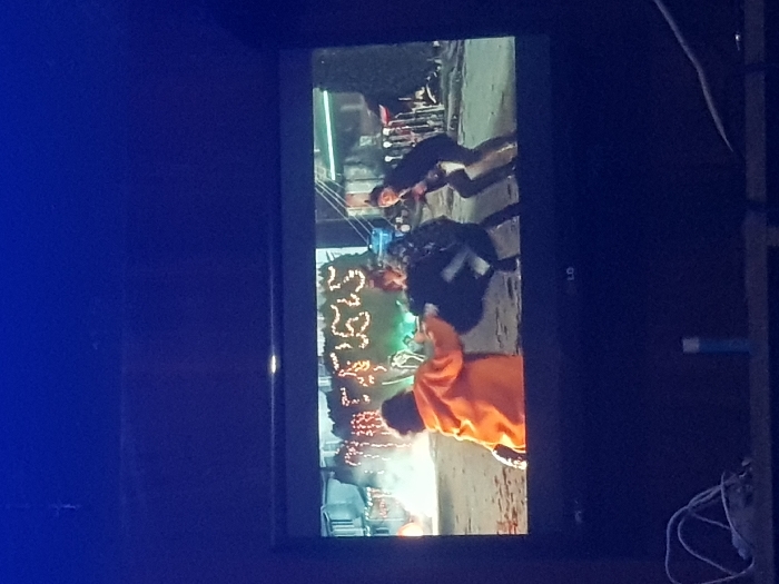 A television displays a scene. Three individuals appear. One is wearing an orange shirt and stands closest to the viewer. Another individual, wearing dark clothing and carrying a backpack, is positioned between the first person and a third individual, who is kneeling and also dressed in dark clothing. In the background, a sign with twinkling lights is visible. The location appears to be outside. The TV's LG logo is visible on its bezel. Cables are seen near the bottom right corner of the device.
