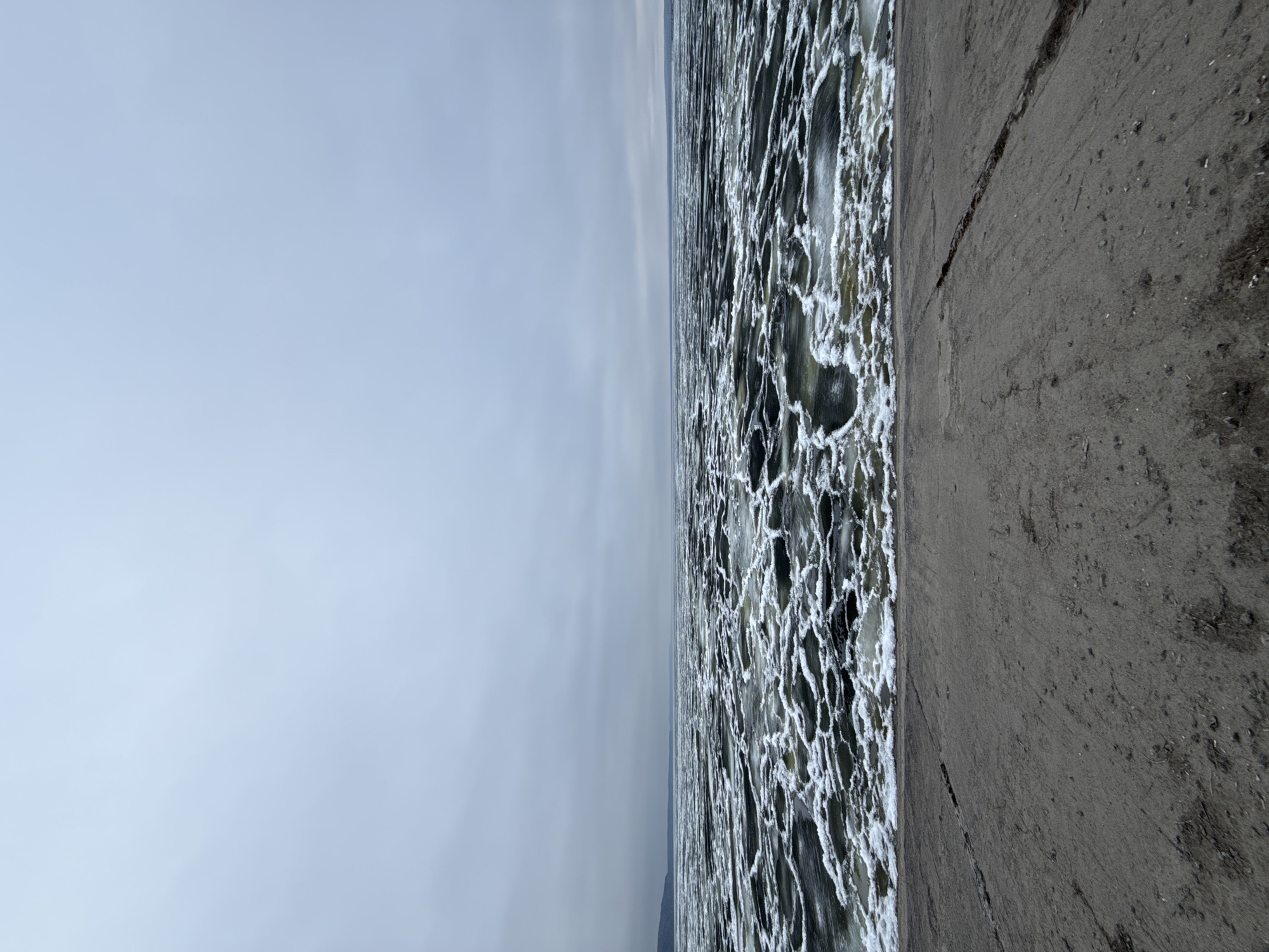 This image captures a winter scene along a body of water, likely a river or lake, near a concrete embankment under an overcast sky. The photograph, oriented vertically, shows a wide expanse of water covered in a fragmented mosaic of ice. White, textured ice floes, possibly pancake ice, intermingle with darker patches of open water, creating an intricate pattern that stretches towards the distant horizon.
On the right side of the image, a grey, rough concrete surface serves as the immediate foreground and embankment. This concrete appears weathered, showing several cracks and scattered debris like small stones and dirt. The left side of the image is dominated by a uniform, pale grey, overcast sky, suggesting a cold, dull winter day.
There are no people or specific activities depicted. The scene conveys a sense of stillness and the raw beauty of a frozen landscape. The ice formations indicate cold weather conditions. Given the context of Chaikovsky, Russia, this is consistent with a view of the Kama River during its winter freeze. No visible text is present in the image.