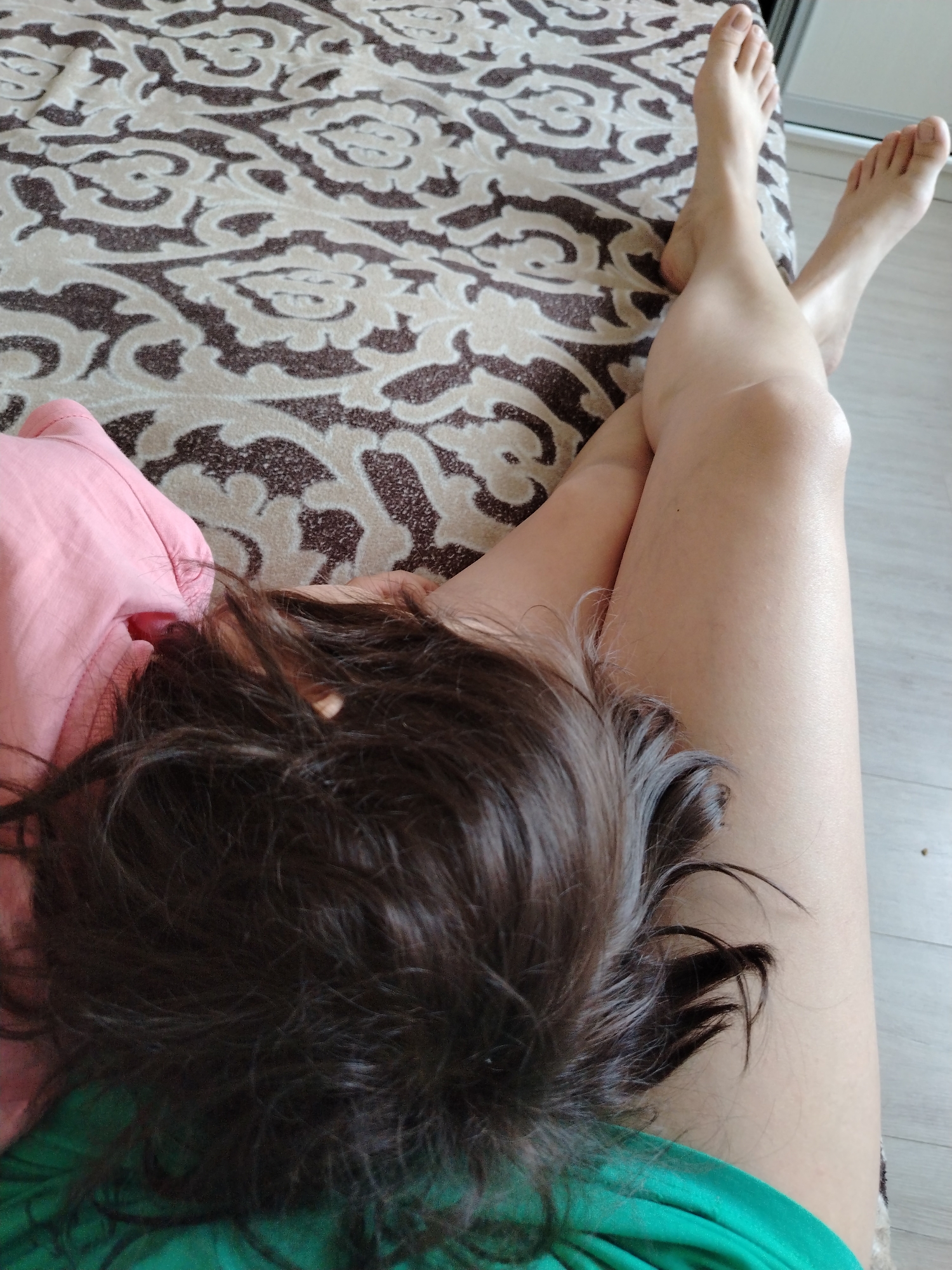A person is lying on a bed with their head resting on their crossed legs. The bed has a patterned beige and brown cover. The person is wearing a pink top on their left and a green top on their right. Their feet are visible at the top of the frame. The floor is a light-colored wood. There is no information provided indicating this image was taken in Noviy, Russia.