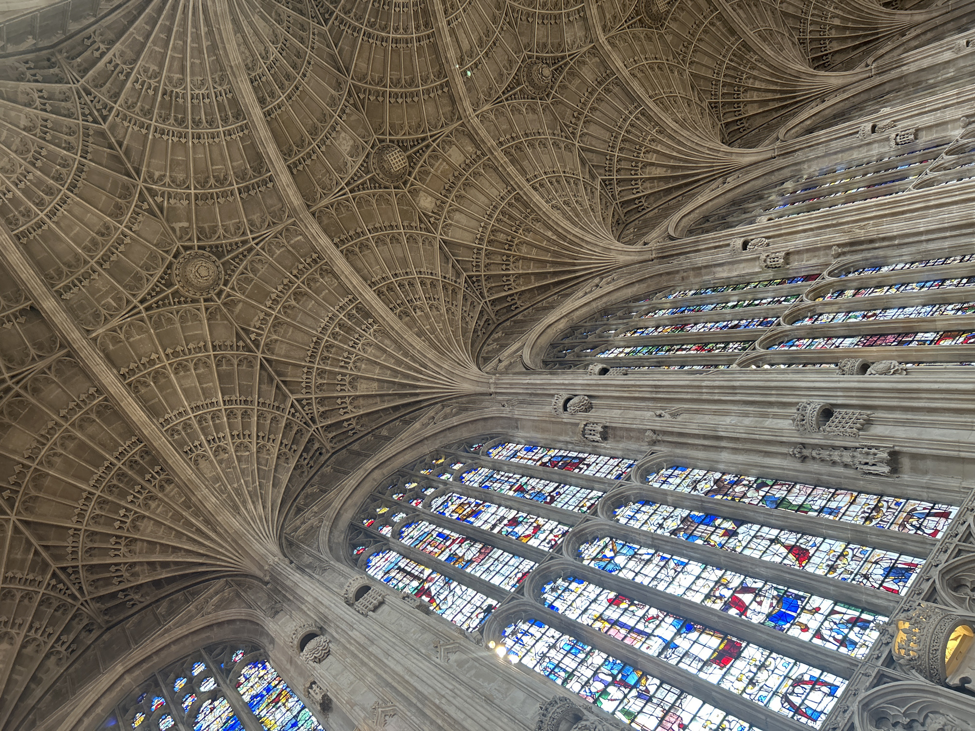 This is a photograph of the fan vaulting ceiling and stained glass windows within King's College Chapel in Cambridge, United Kingdom. The image is taken from a low angle, looking upwards.
The ceiling features intricate stone carvings in a fan vaulting pattern, with multiple ribs extending from points on the walls and converging towards the apex. The stone appears to be a light, weathered color. Within the complex pattern of the vaulting, there are circular decorative elements.
The walls are dominated by tall, arched stained glass windows. The windows are filled with vibrant colored glass depicting various scenes and figures, with dominant colors including blue, red, yellow, and green. The glass is divided into numerous panes.
In the lower parts of the image, glimpses of ornate stone tracery and architectural details, such as small carved figures and decorative crowns, are visible around the window frames. The overall impression is one of grand Gothic architecture. There are no people visible in the image.