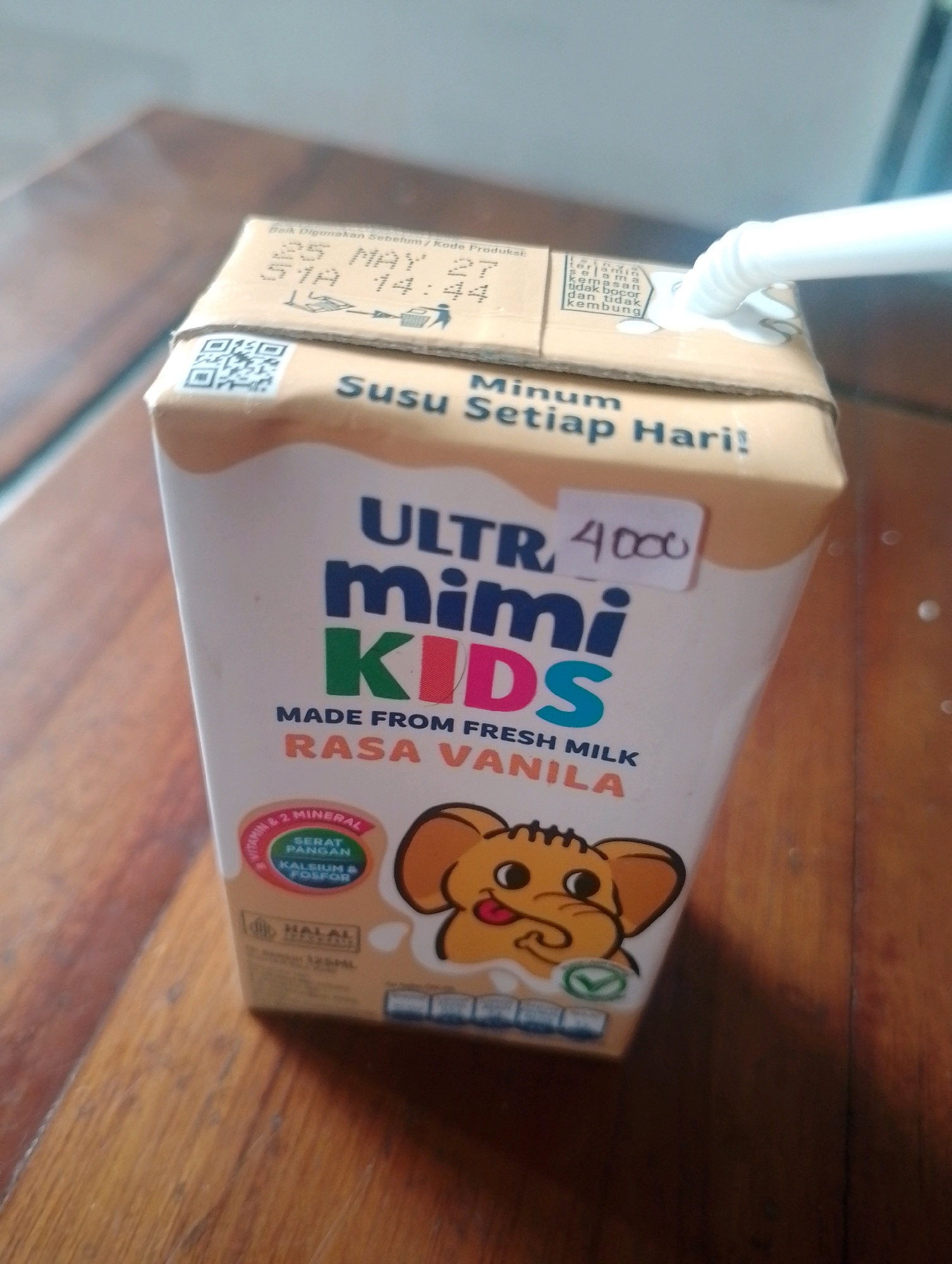 The image displays a carton of UHT milk on a wooden surface, likely a table. The carton is light brown and features bold blue and multi-colored text advertising "ULTRA mimi KIDS" and "MADE FROM FRESH MILK RASA VANILA". A sticker with "4000" is affixed to the carton, possibly indicating a price. A white straw extends from the top of the carton. The carton has a QR code on one side and nutritional information, including "VITAMIN & 2 MINERAL" and "HALAL" certification, on another. The wooden surface appears to be a typical indoor setting. There is no visible indication of the city of Kendalrejo, Indonesia, or any specific location details beyond what is printed on the product packaging. The image is taken from a slightly elevated angle, with the carton positioned in the center.